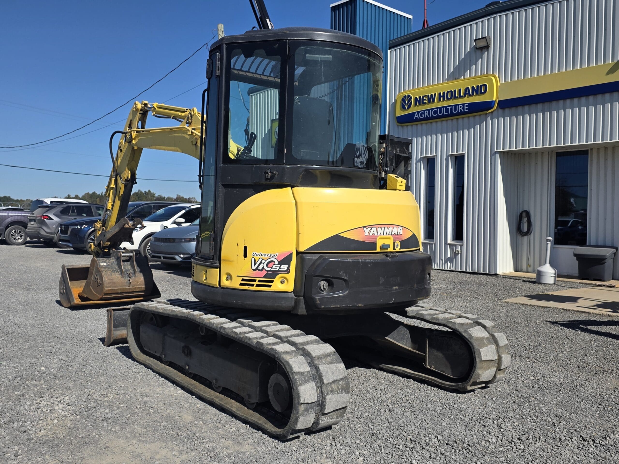 Une mini-pelle Yanmar jaune est garée sur du gravier devant un bâtiment de New Holland Agriculture ; d'autres véhicules et engins sont visibles à l'arrière-plan.