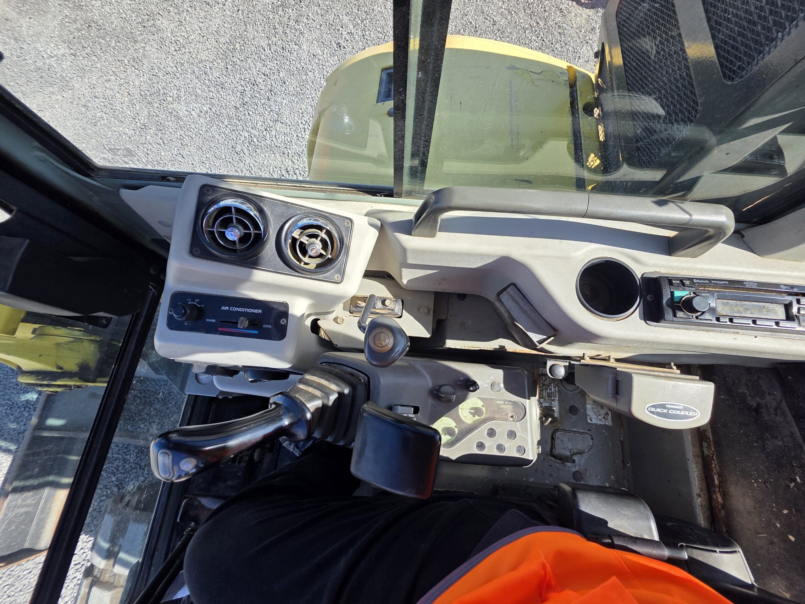 Vue intérieure de la cabine d'un engin de chantier, montrant le tableau de bord, le joystick, les pédales et une partie d'une personne portant un gilet orange, avec la lumière du soleil qui pénètre par la vitre.