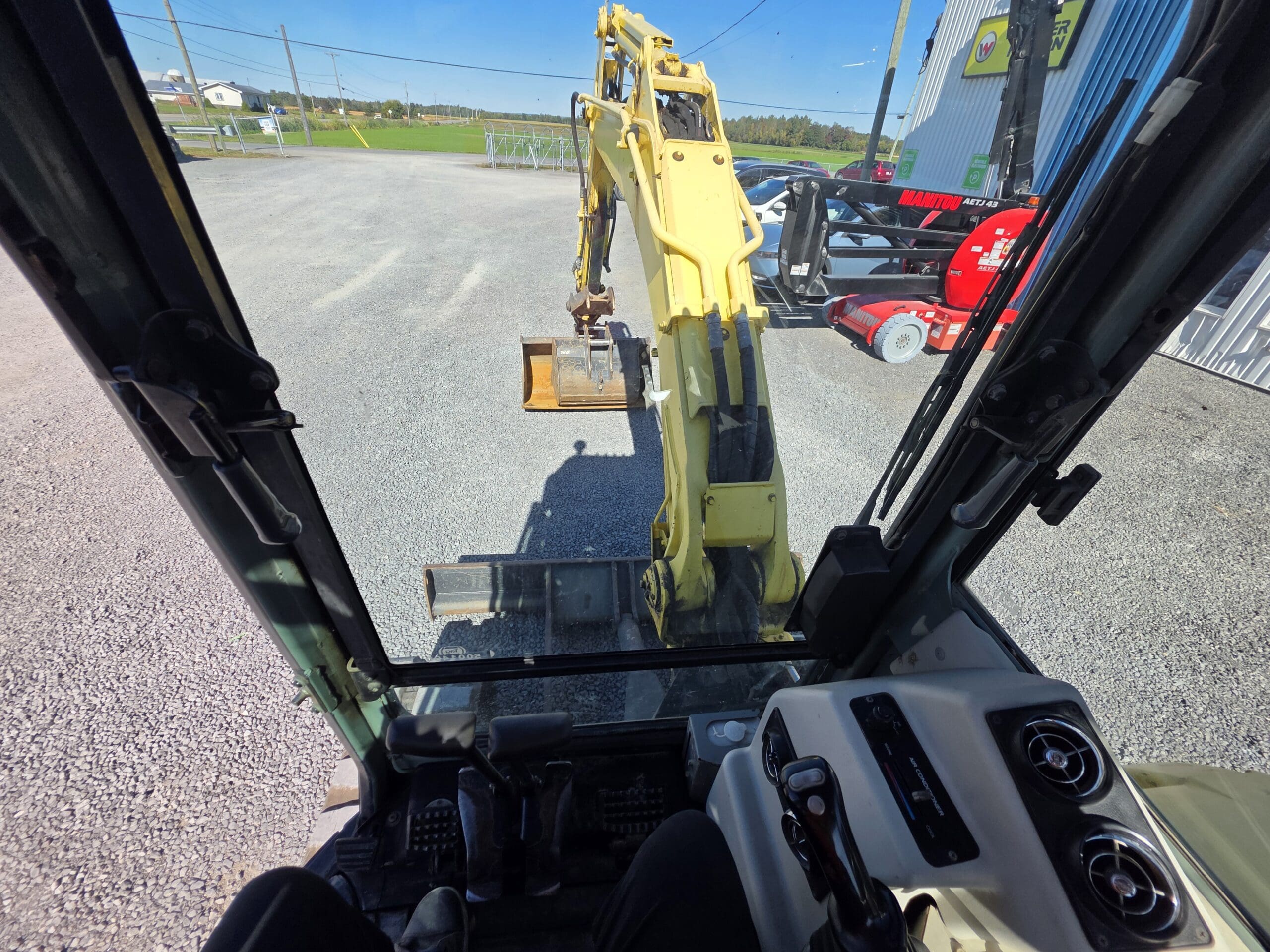 Vue depuis l'intérieur de la cabine d'un engin de chantier, montrant le bras jaune d'une pelleteuse au-dessus du gravier. On aperçoit du matériel et des bâtiments à l'arrière-plan.