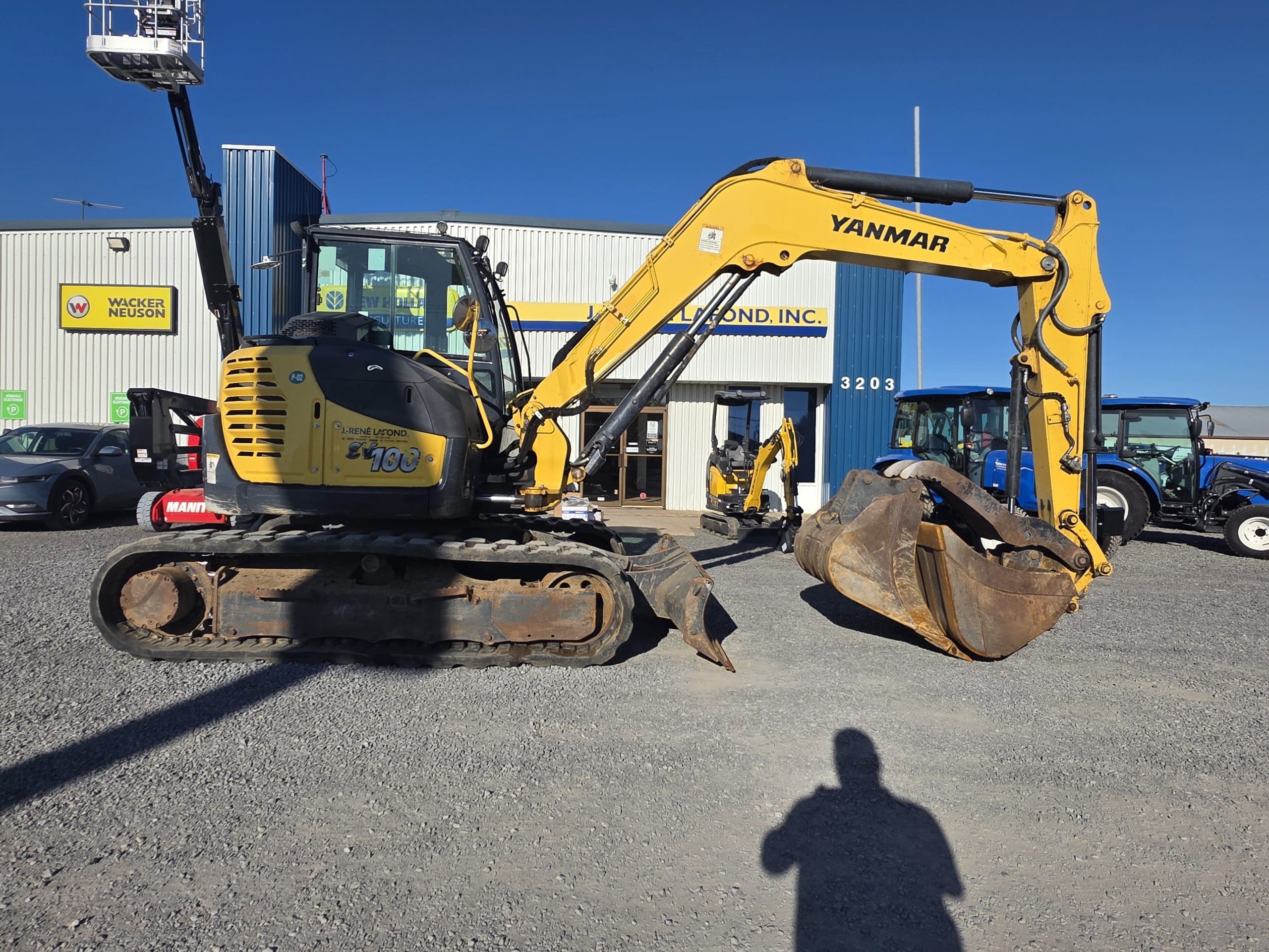 Une pelleteuse Yanmar jaune est garée sur du gravier devant un bâtiment bleu et blanc, par une journée ensoleillée. On aperçoit des véhicules et du matériel à l'arrière-plan, tandis qu'une ombre humaine se détache au premier plan.