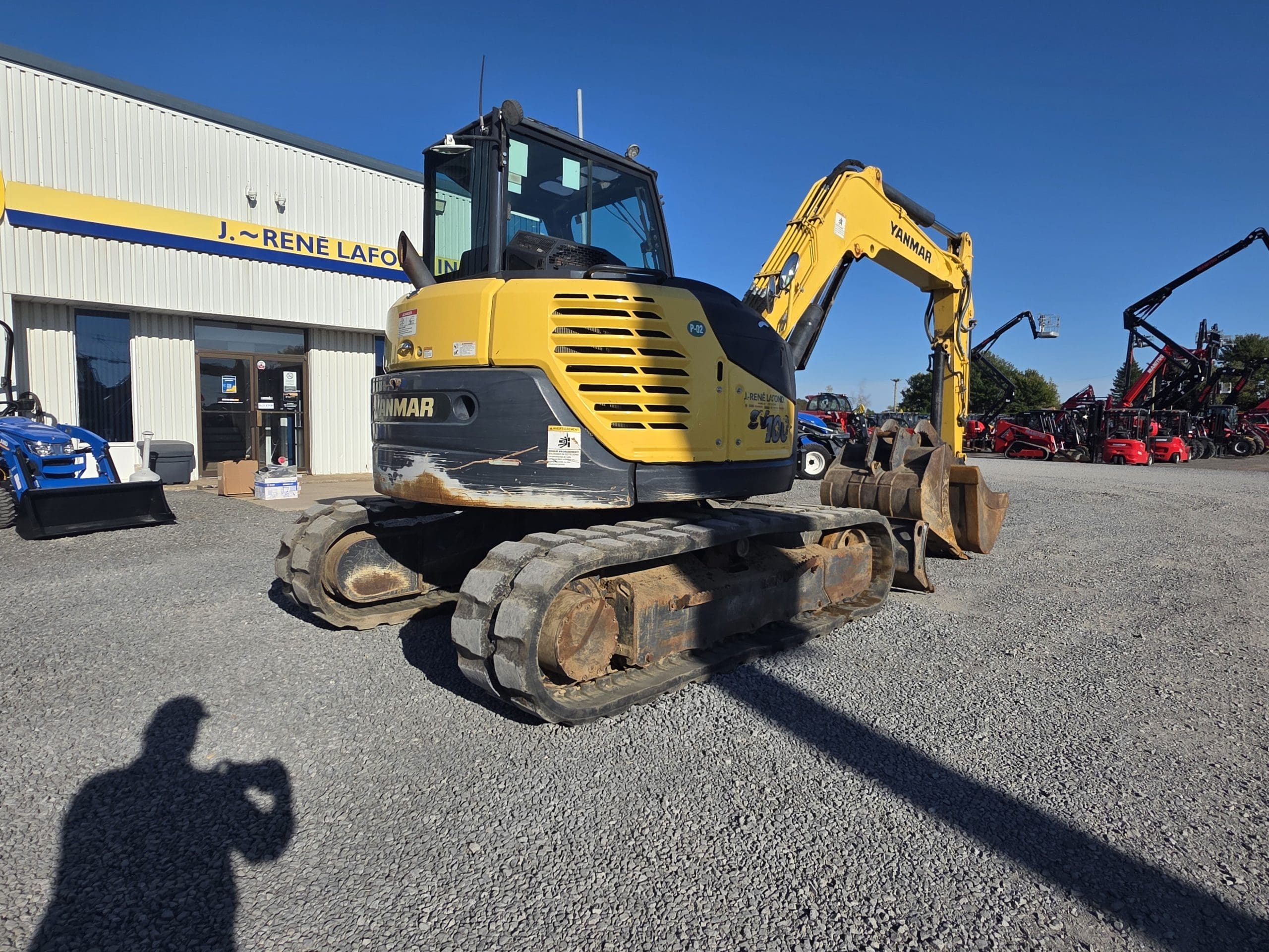 Une pelleteuse Yanmar jaune est garée sur du gravier devant un bâtiment. D'autres engins sont visibles à l'arrière-plan, et l'ombre d'une personne se dessine au premier plan.