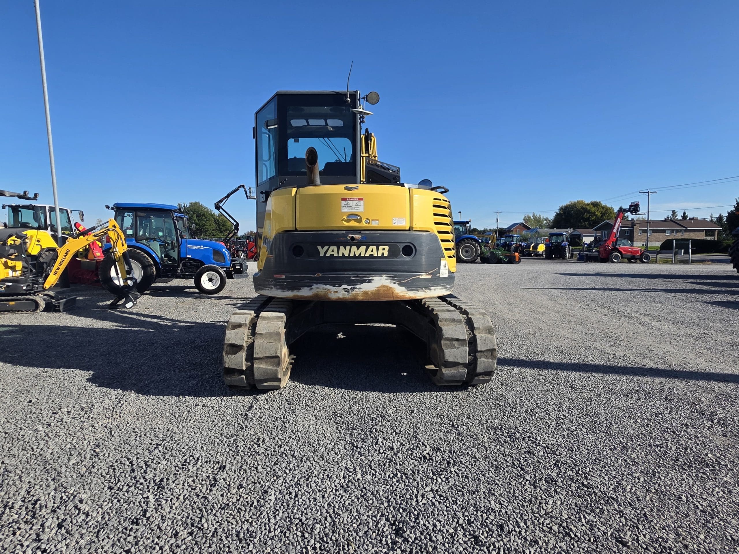 Une pelleteuse Yanmar jaune est garée sur un terrain en gravier sous un ciel bleu limpide, avec d'autres engins de chantier et véhicules visibles à l'arrière-plan.