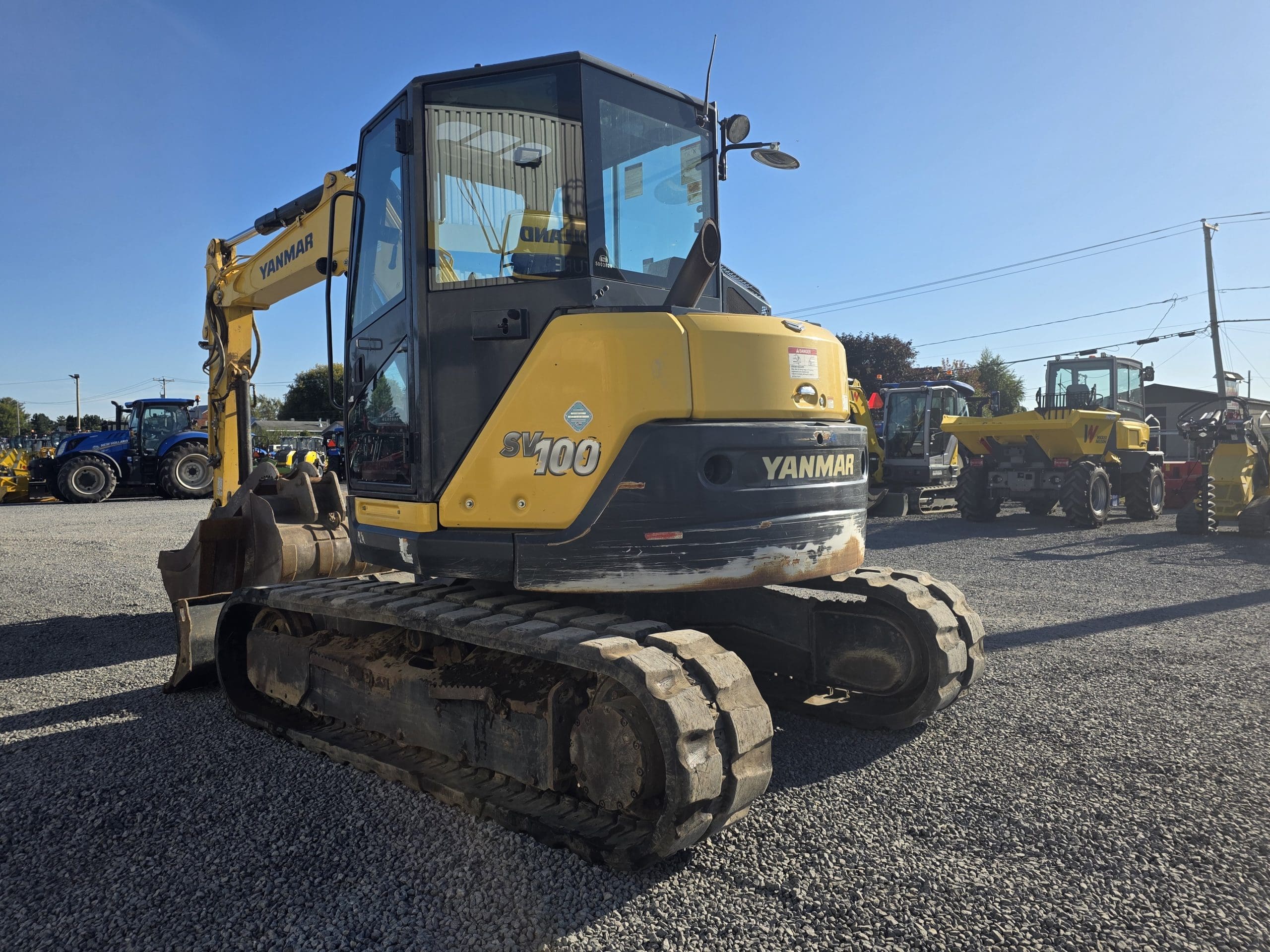 Une pelleteuse Yanmar SV100 jaune est garée sur du gravier ; on aperçoit d'autres engins de chantier à l'arrière-plan, sous un ciel bleu limpide.