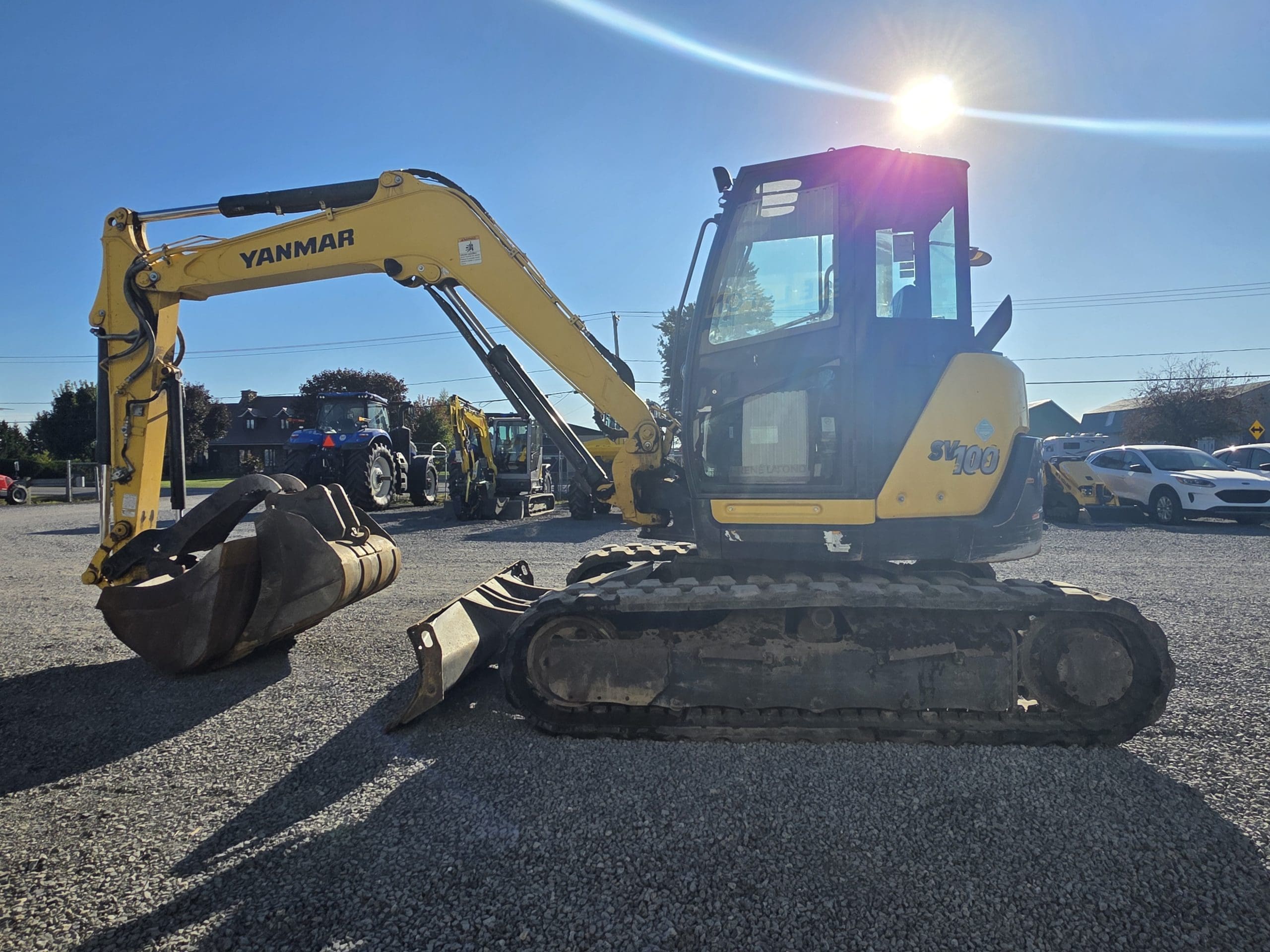 Une pelleteuse Yanmar jaune est garée sur du gravier dans un terrain, sous un soleil radieux, avec des voitures et des arbres en arrière-plan.