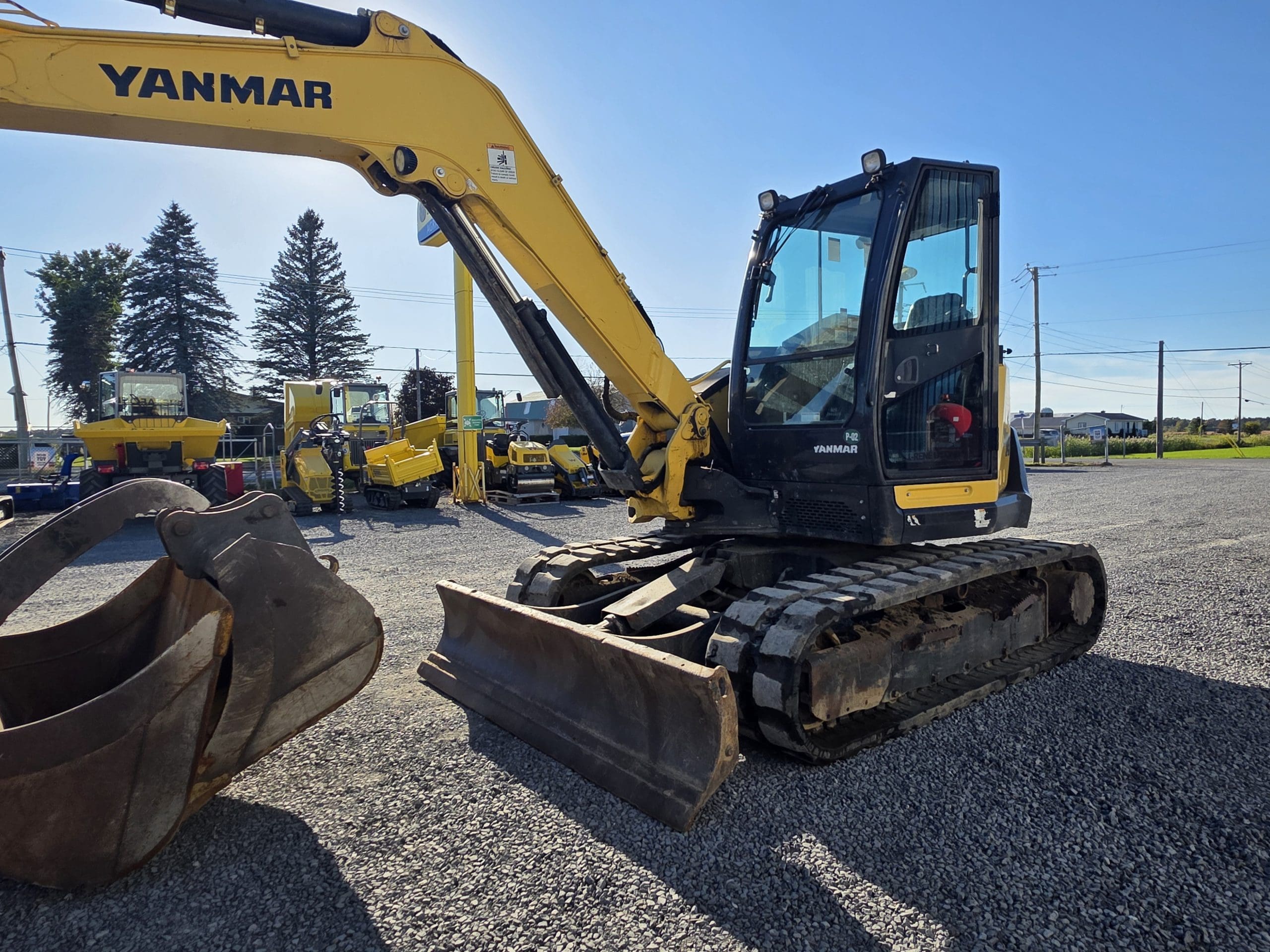 Une pelleteuse Yanmar jaune équipée d'une lame avant est garée sur du gravier, avec d'autres engins de chantier en arrière-plan, sous un ciel bleu limpide.