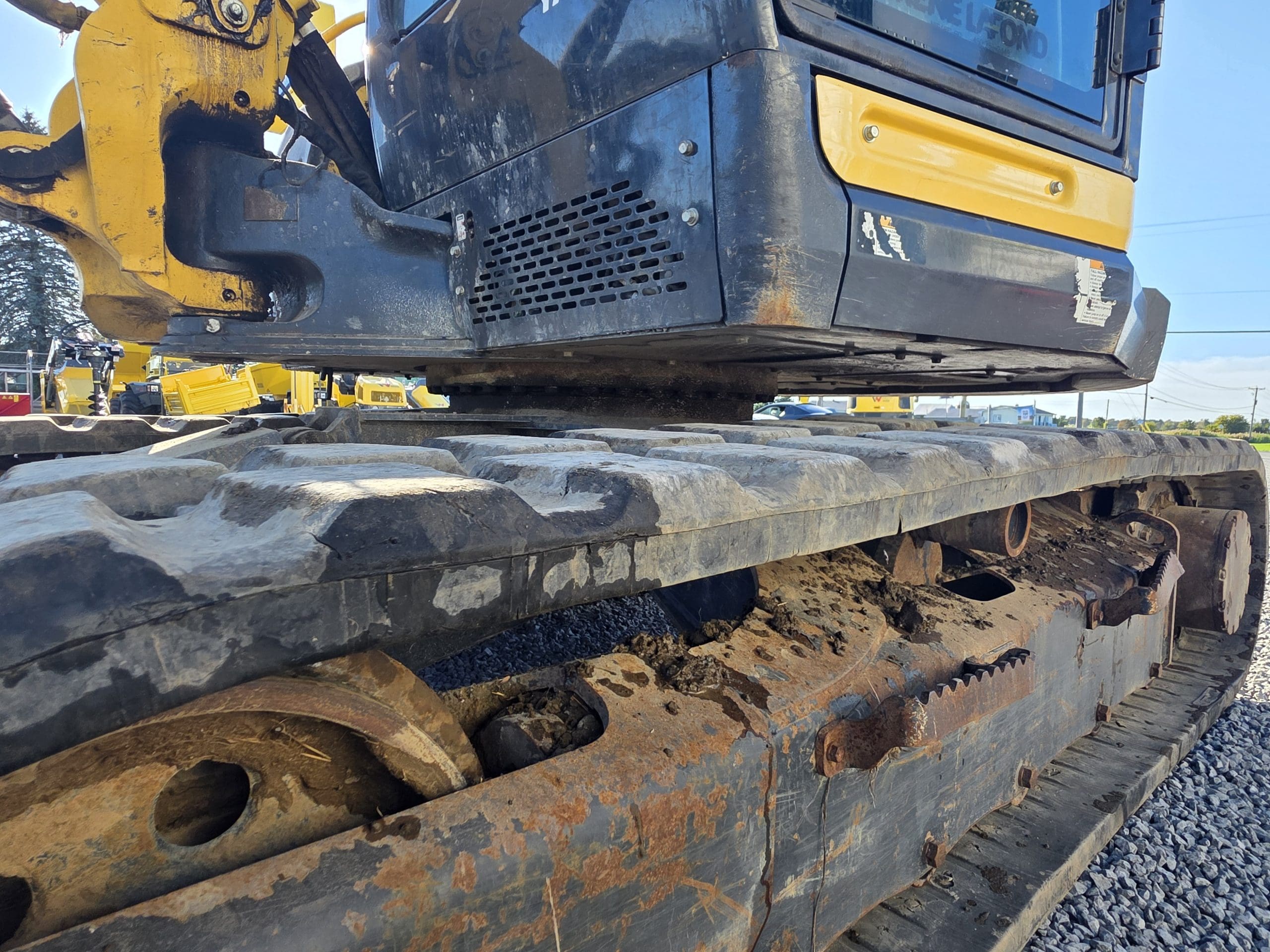 Gros plan sur les chenilles métalliques et le train de roulement d'une pelleteuse jaune et noire, présentant des traces de rouille et de saleté. La machine est posée sur du gravier, avec d'autres engins de chantier visibles à l'arrière-plan.