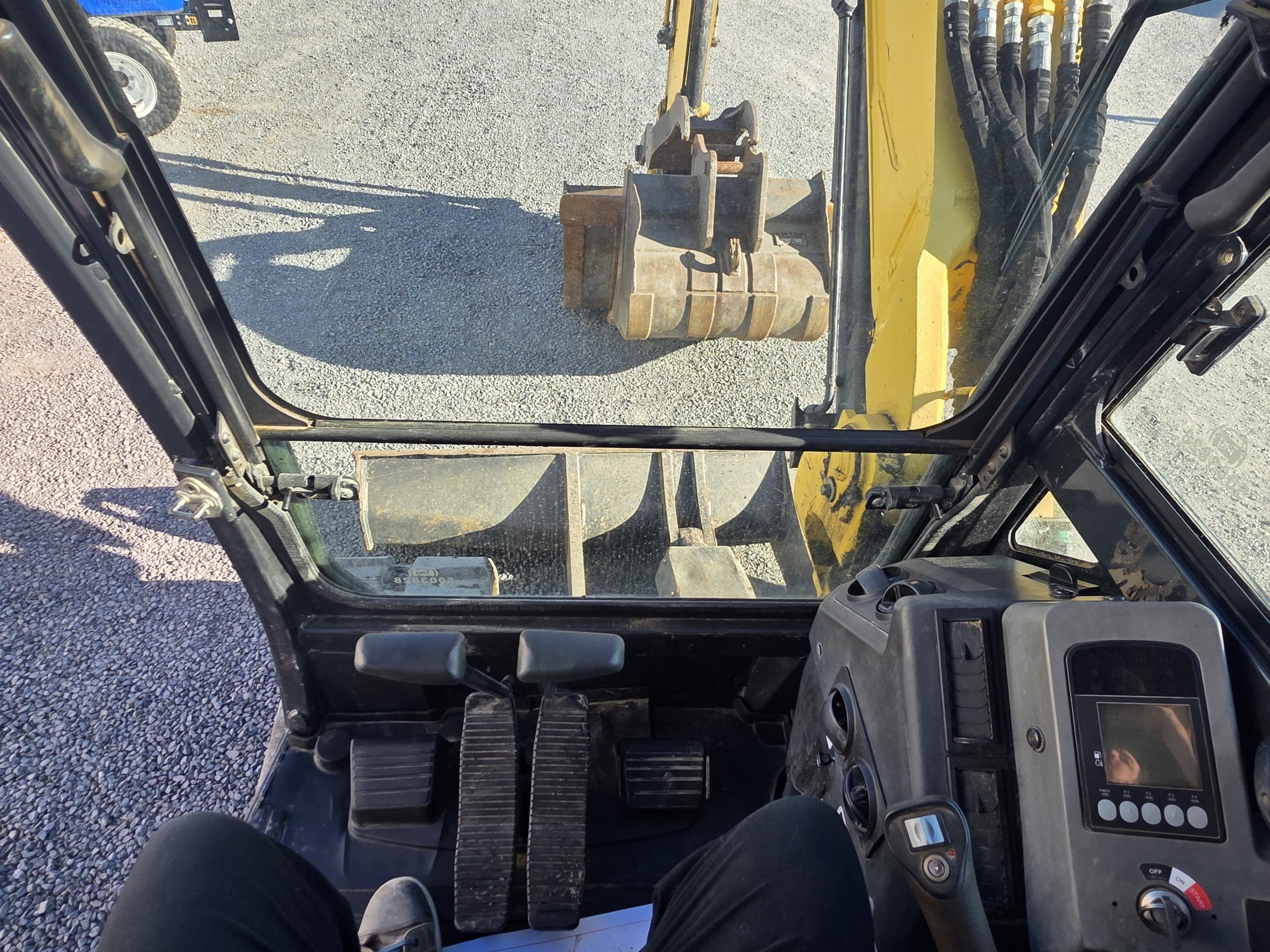 Vue depuis l'intérieur de la cabine d'une pelleteuse, montrant les pédales, les commandes et un écran numérique ; on aperçoit, à travers le pare-brise, le bras et le godet jaunes de la pelleteuse sur une surface de gravier.