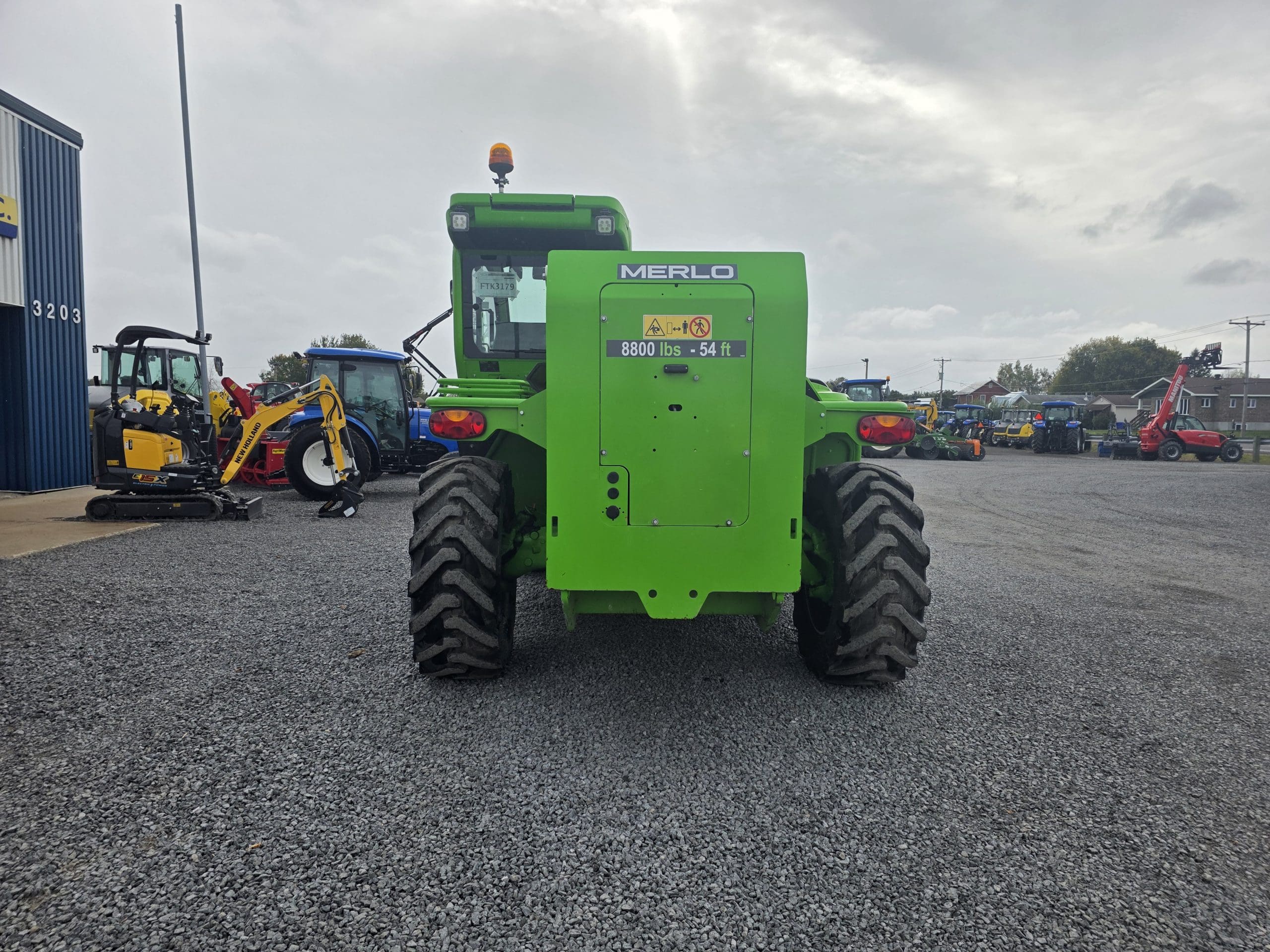 Un chariot télescopique Merlo vert vif est garé sur du gravier, vu de l'arrière. Divers engins et équipements de chantier sont visibles à l'arrière-plan, près d'un bâtiment bleu, sous un ciel nuageux.