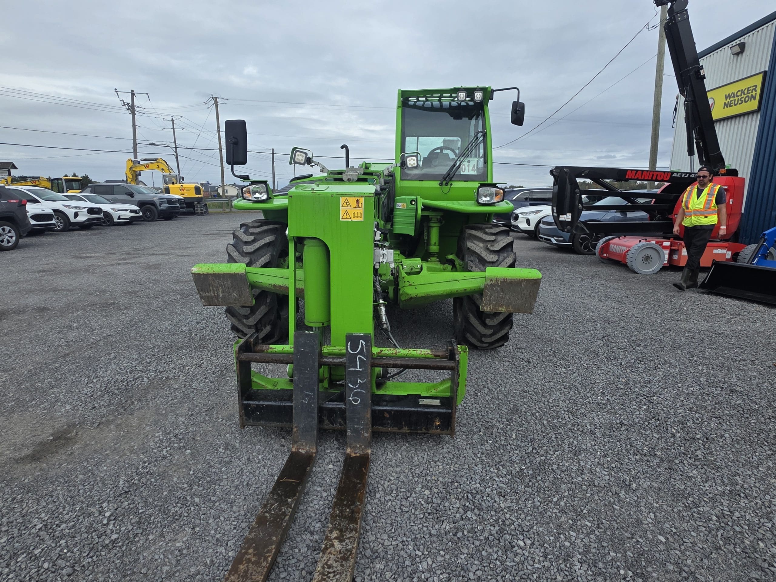 Un chariot élévateur vert vif garé sur du gravier, face à la caméra, avec des voitures et des engins de chantier en arrière-plan, ainsi qu'un ouvrier portant un gilet de sécurité qui se tient à proximité.
