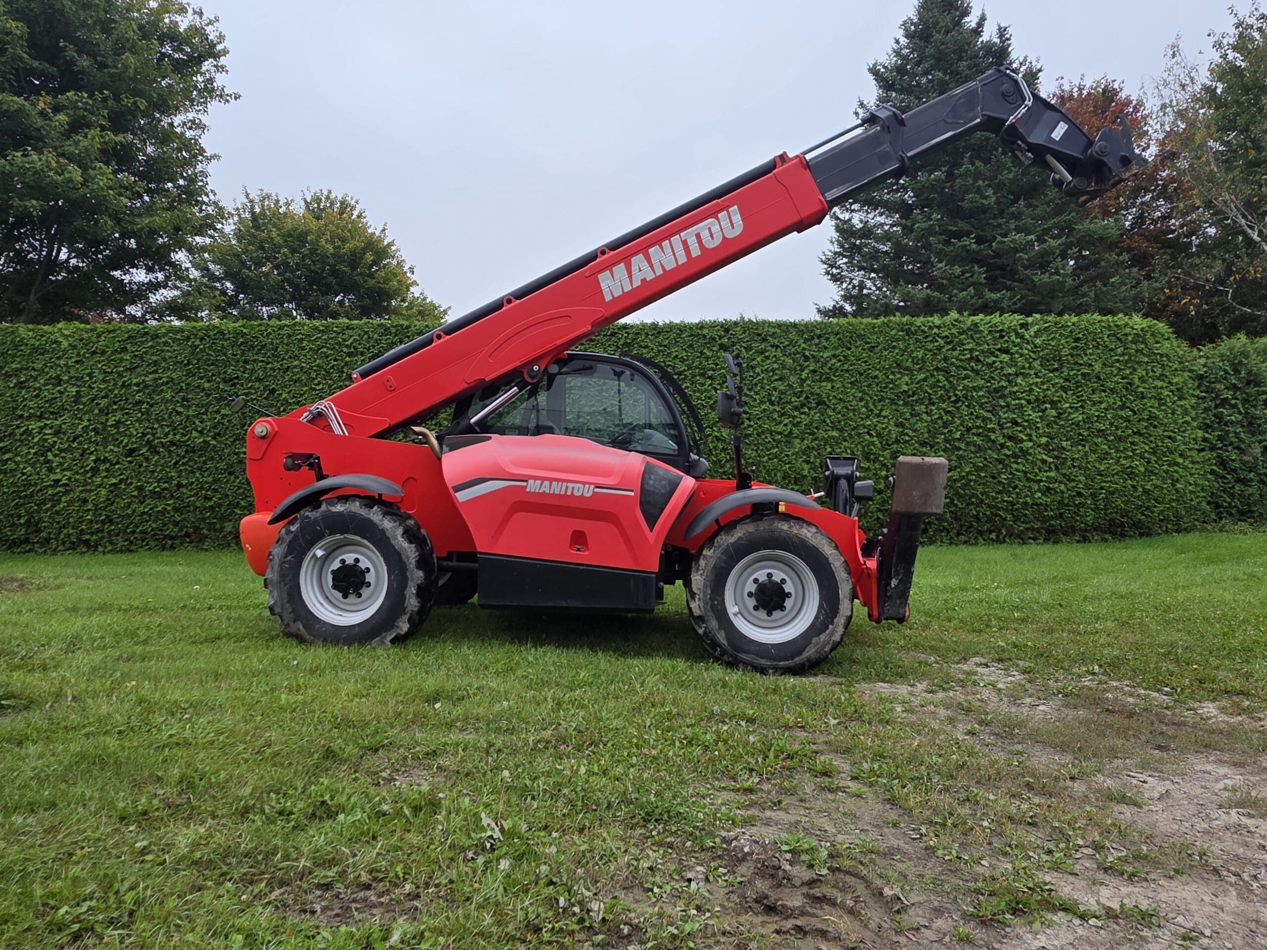 Un chariot télescopique Manitou rouge, équipé d'une flèche allongée, est garé sur une pelouse verte, avec une haute haie et des arbres en arrière-plan.