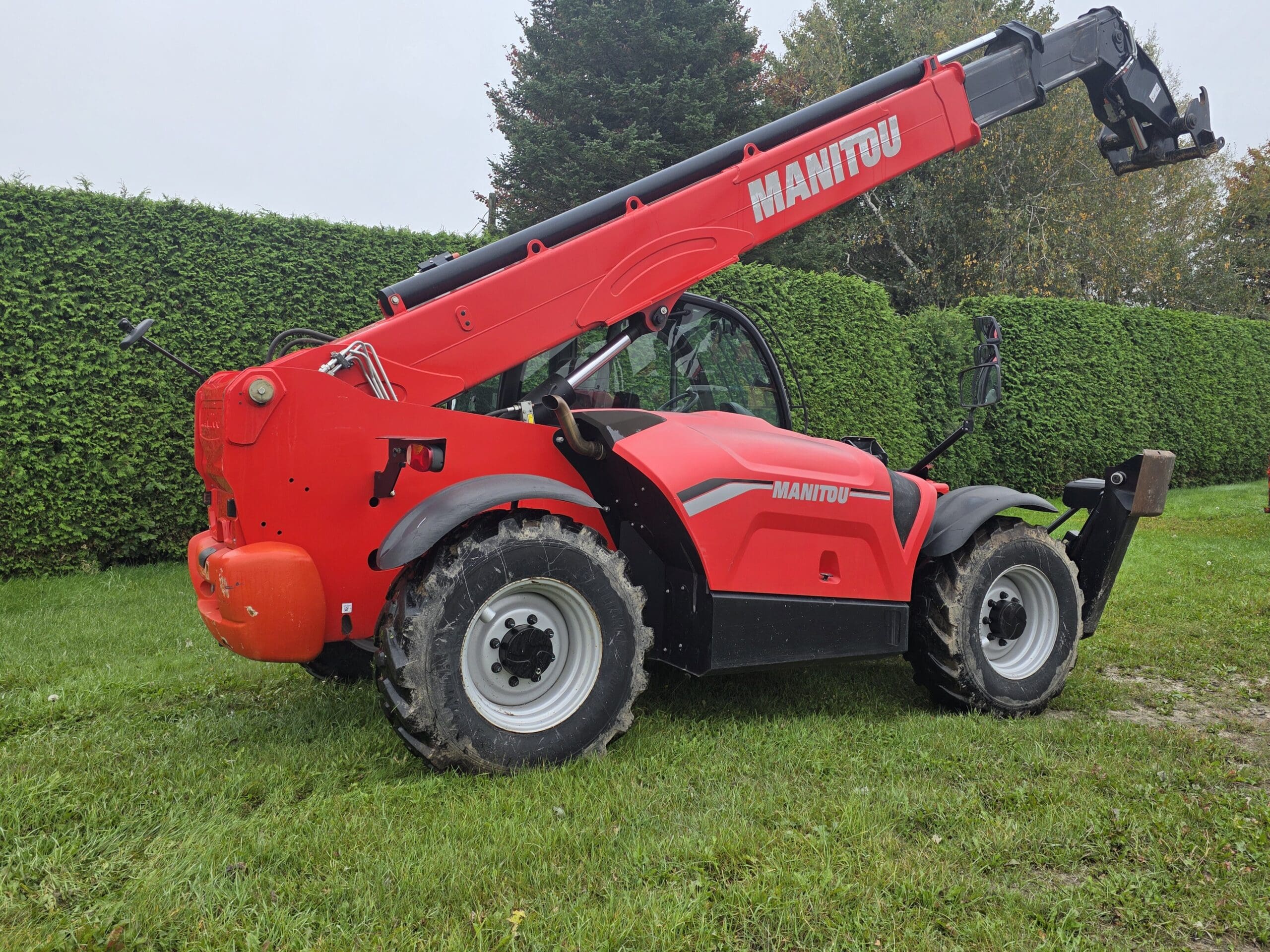 Un chariot télescopique Manitou rouge est garé sur l'herbe, près de hautes haies vertes et d'arbres, par une journée nuageuse.