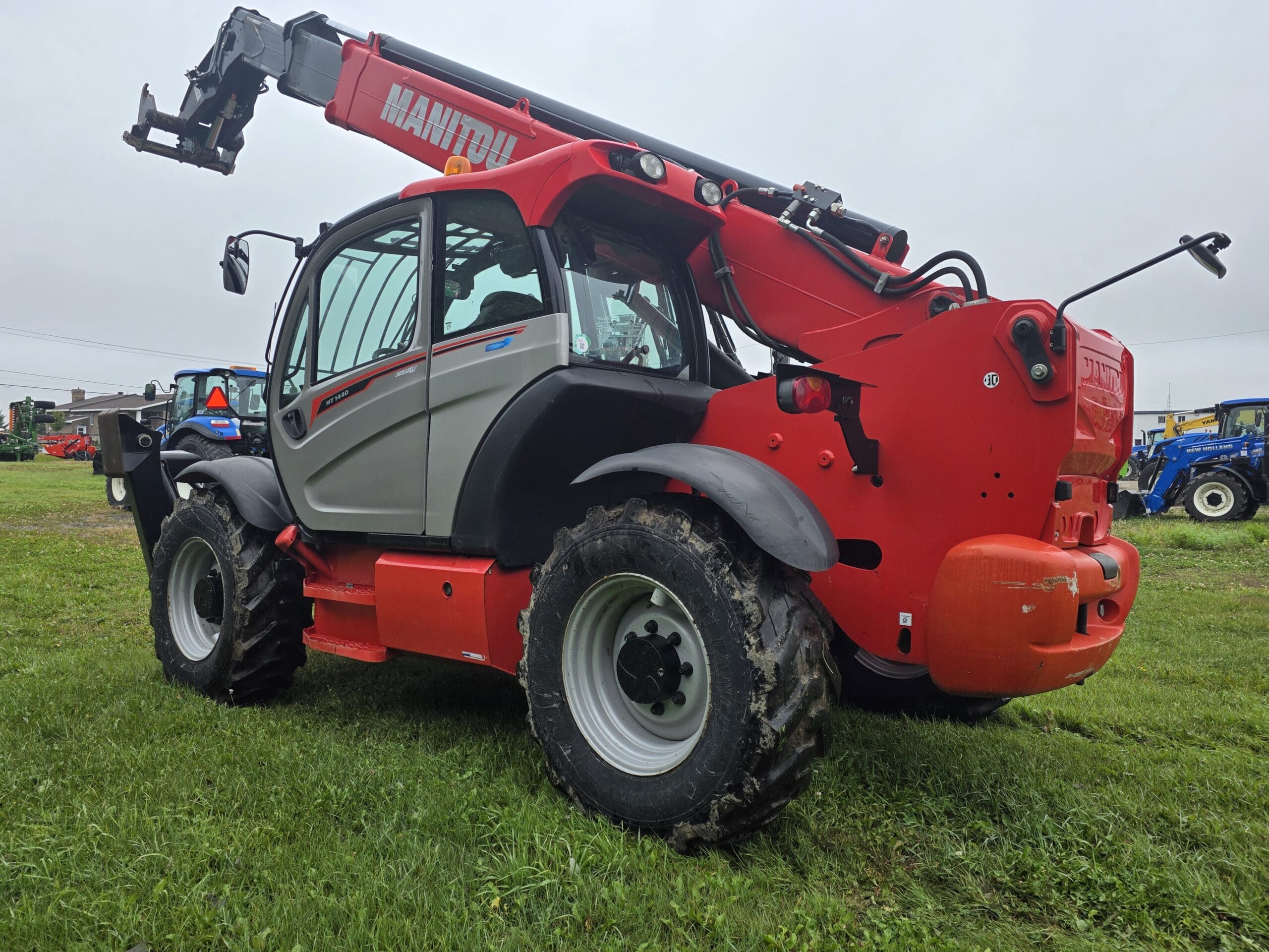Un chariot télescopique Manitou rouge, équipé de gros pneus et d'une flèche allongée, est garé sur un terrain herbeux en plein air ; on aperçoit d'autres machines agricoles à l'arrière-plan, sous un ciel nuageux.