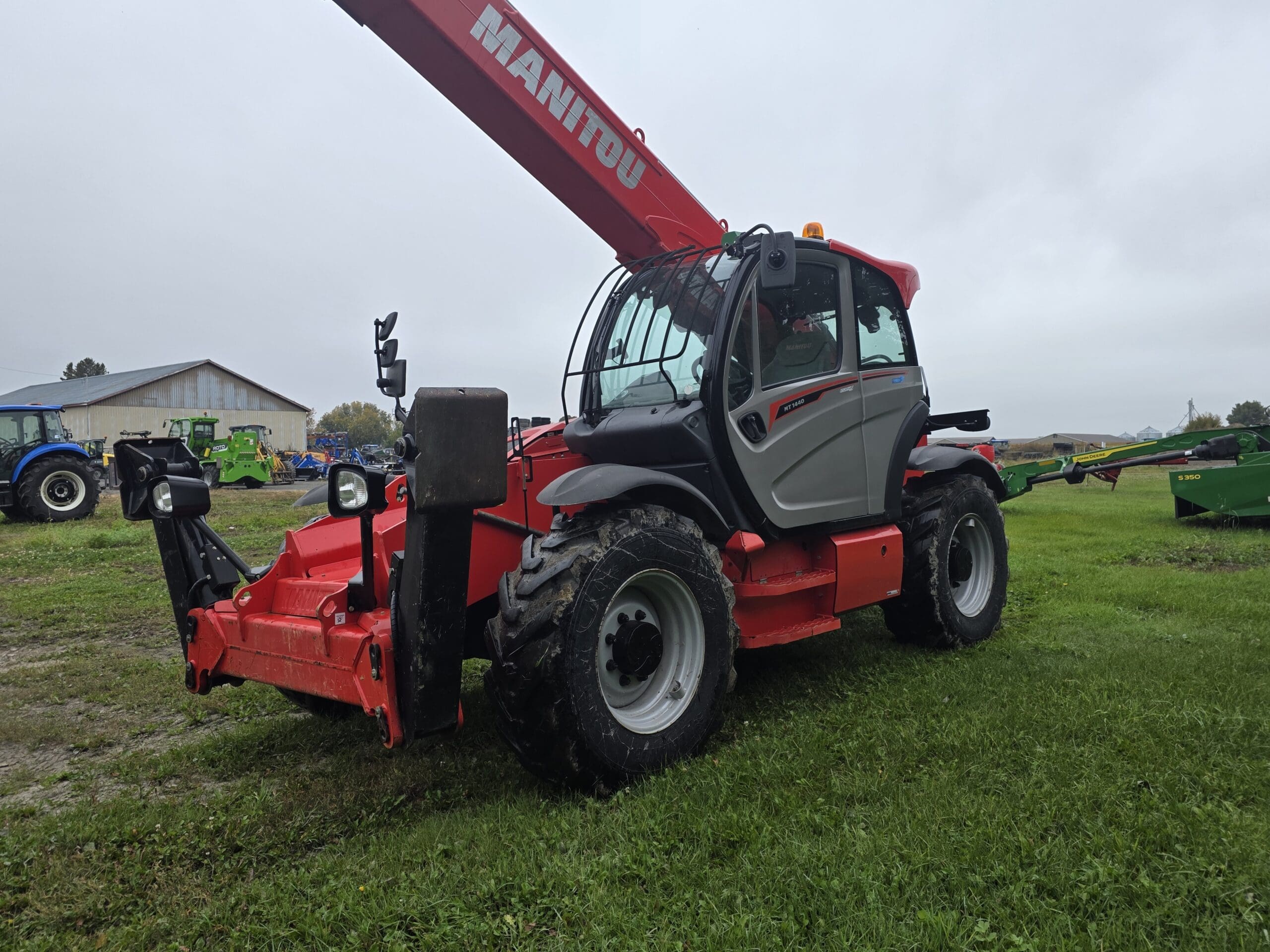 Un chariot télescopique Manitou rouge, équipé de gros pneus, est garé sur l'herbe près des bâtiments agricoles et d'autres machines, sous un ciel nuageux.