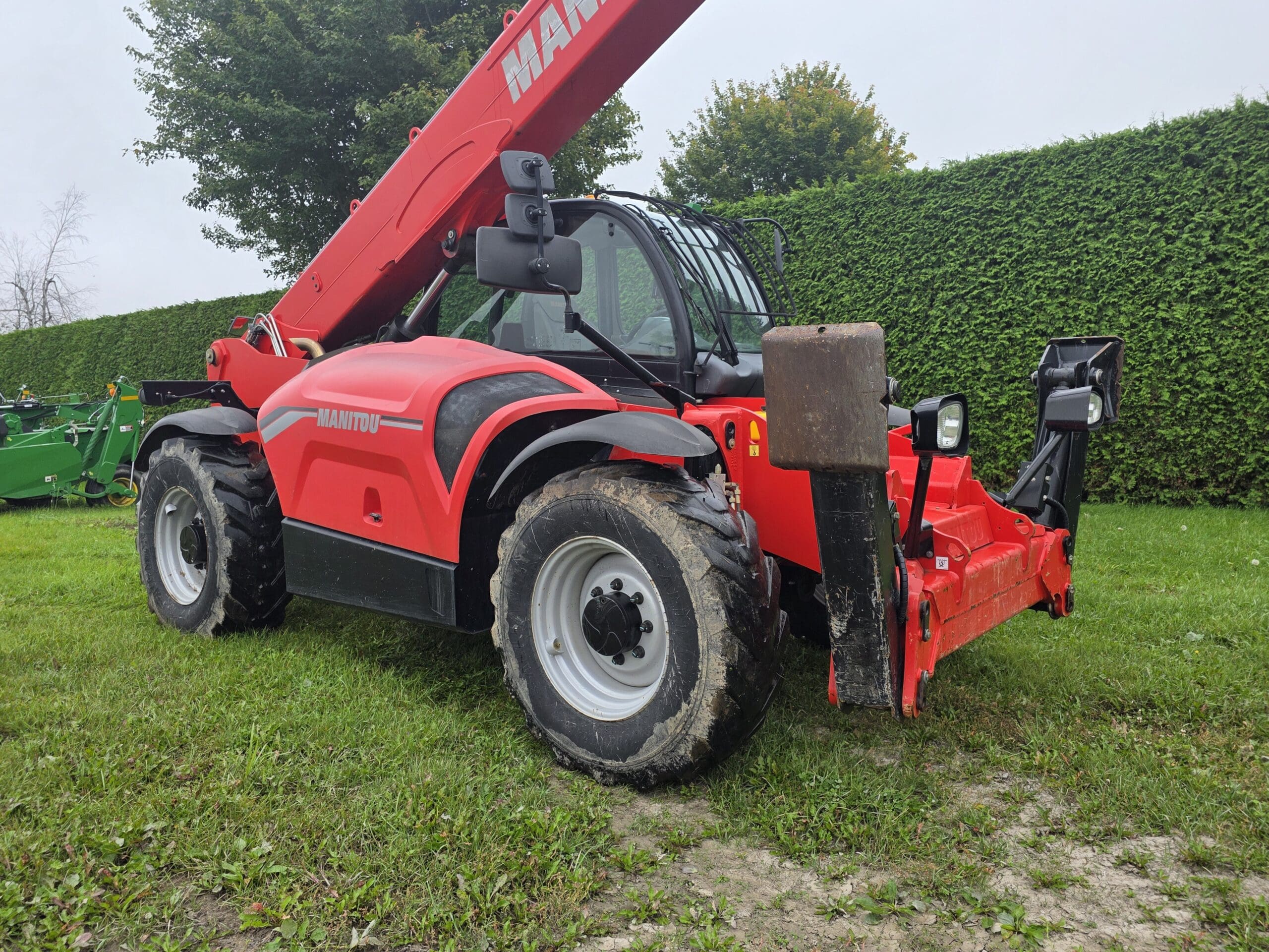 Un chariot télescopique Manitou rouge équipé de gros pneus est garé sur l'herbe. On aperçoit des arbres et une haute haie verte en arrière-plan. La flèche est relevée et la machine est légèrement boueuse.