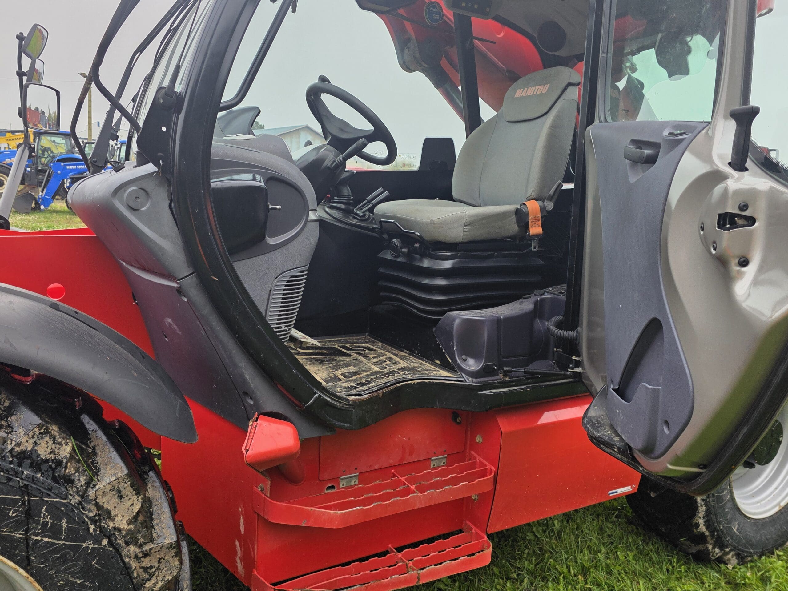 Gros plan sur une porte ouverte dévoilant l'habitacle d'un tracteur rouge, où l'on distingue le siège du conducteur, le volant, les commandes et les marchepieds boueux. Le tracteur est garé sur l'herbe.
