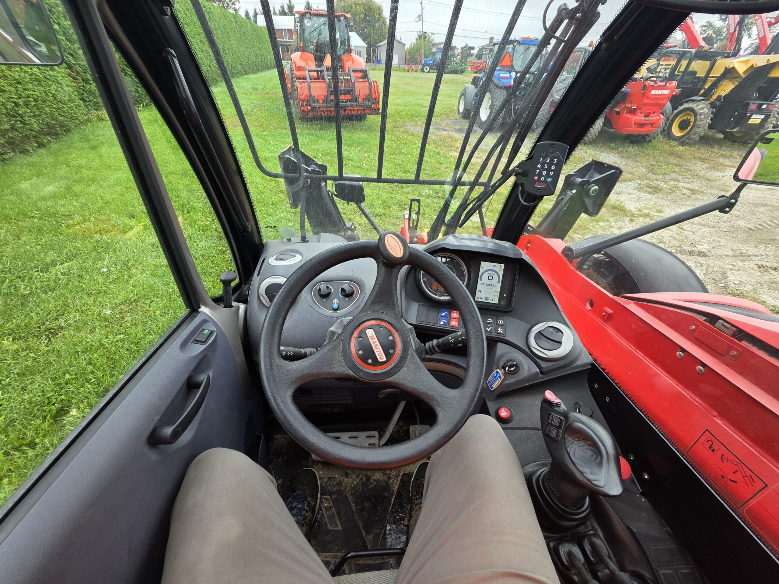 Vue depuis l'intérieur de la cabine d'un tracteur rouge, montrant le volant, les commandes du tableau de bord et les jambes du conducteur. D'autres tracteurs et engins sont visibles à l'extérieur à travers le pare-brise.