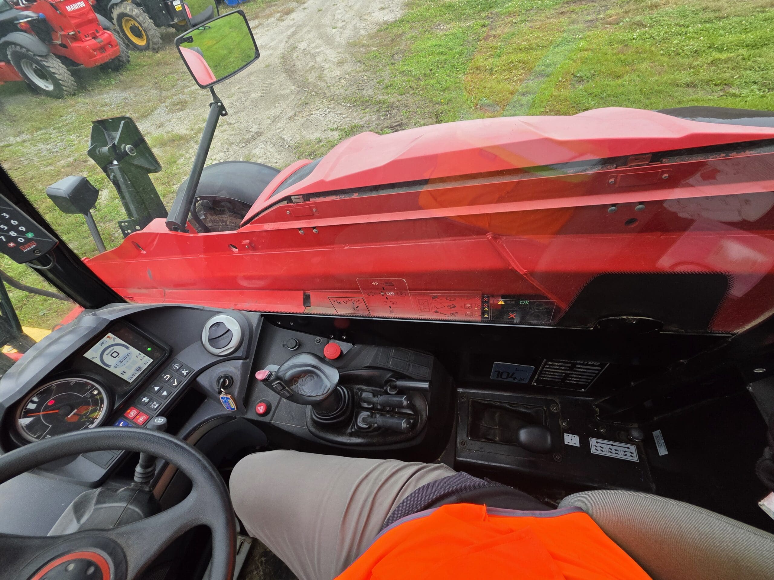 Vue depuis l'intérieur de la cabine d'un tracteur rouge, montrant le volant, le tableau de bord, les leviers et une partie d'une personne vêtue d'un pantalon gris et d'une chemise orange, assise au poste de conduite. À l'extérieur, on aperçoit de l'herbe et des machines.