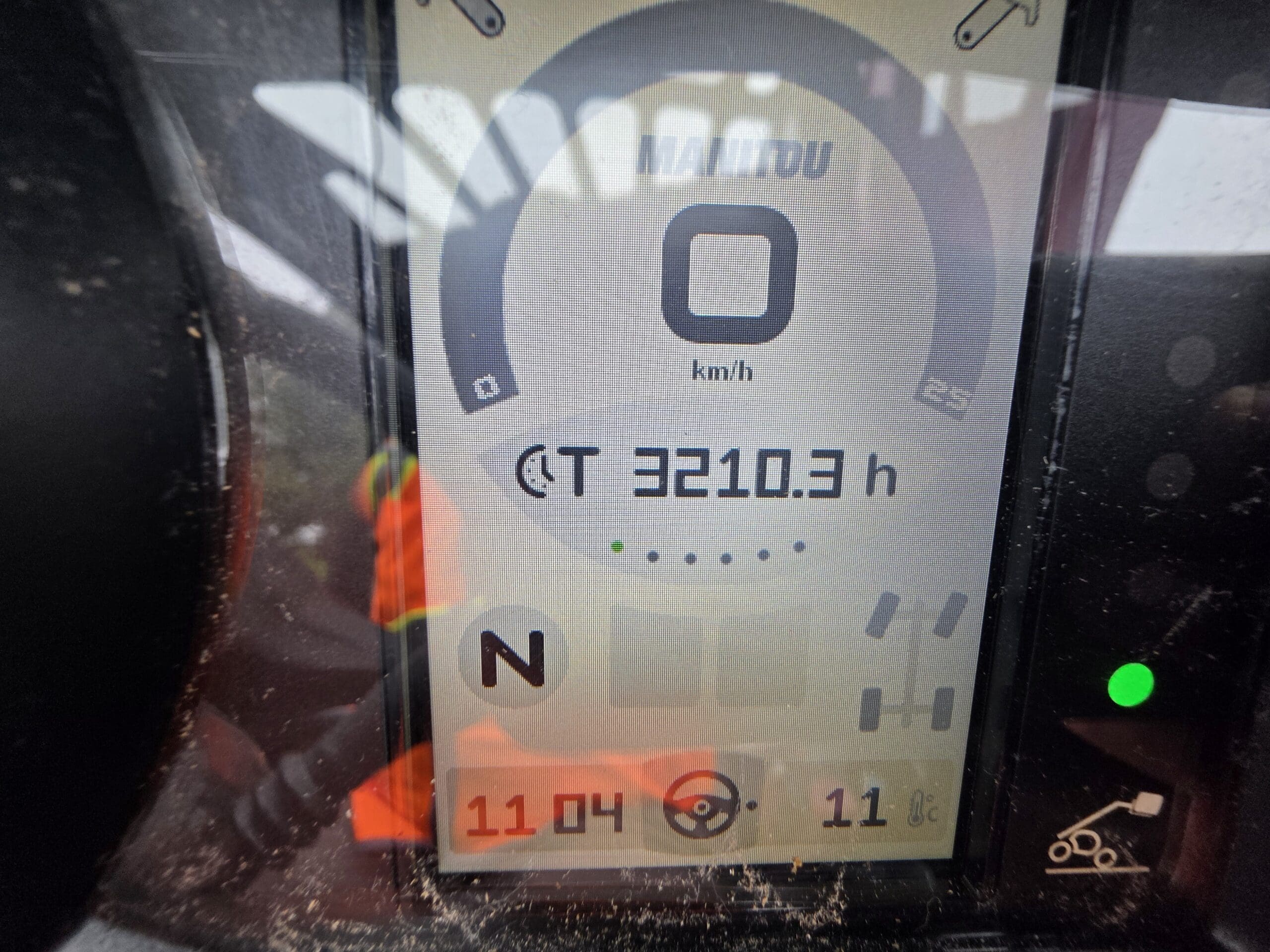 Un écran numérique affiche une vitesse de 0 km/h, 3 210,3 heures de fonctionnement, une boussole orientée vers le nord, des jauges, ainsi que les chiffres 11 h 04 et 11 °C. Le reflet d'une personne portant un gilet de sécurité est visible sur l'écran.