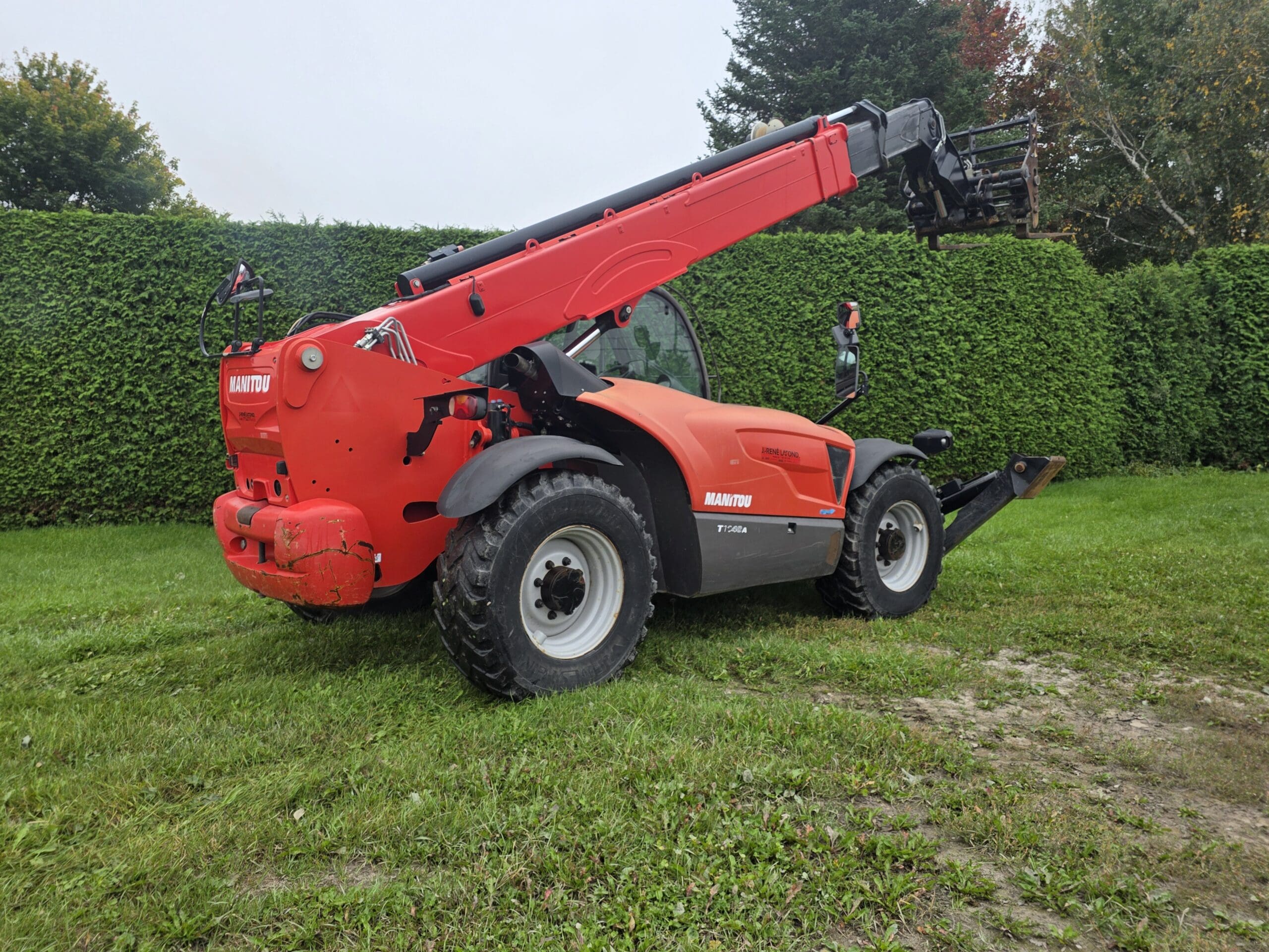 Un chariot télescopique Manitou rouge, équipé d'une flèche allongée, est garé sur l'herbe près d'une haute haie verte, avec quelques arbres en arrière-plan.