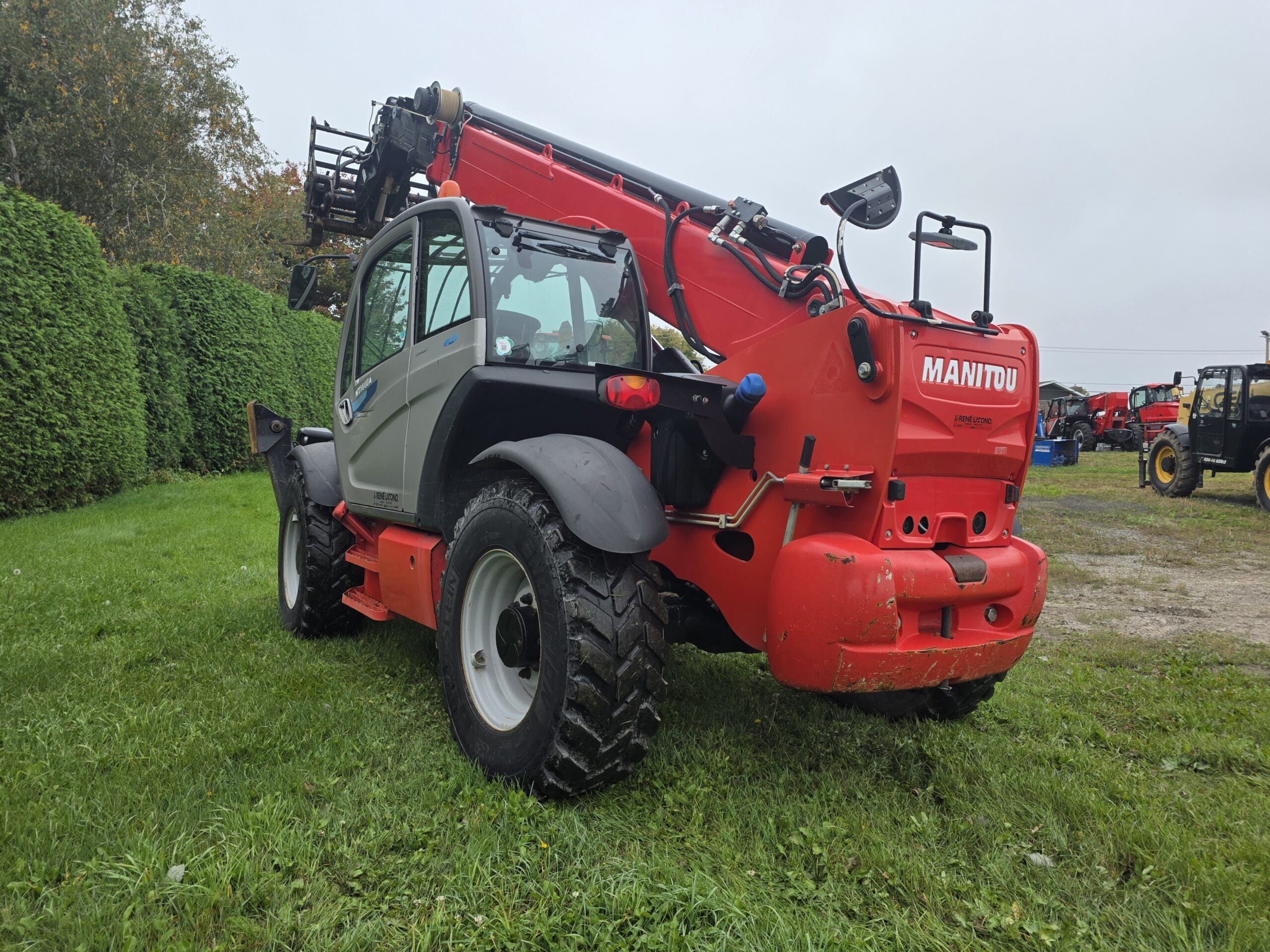 Un chariot télescopique Manitou rouge est garé sur l'herbe près de buissons, sa flèche légèrement relevée. D'autres véhicules sont visibles à l'arrière-plan par une journée nuageuse.