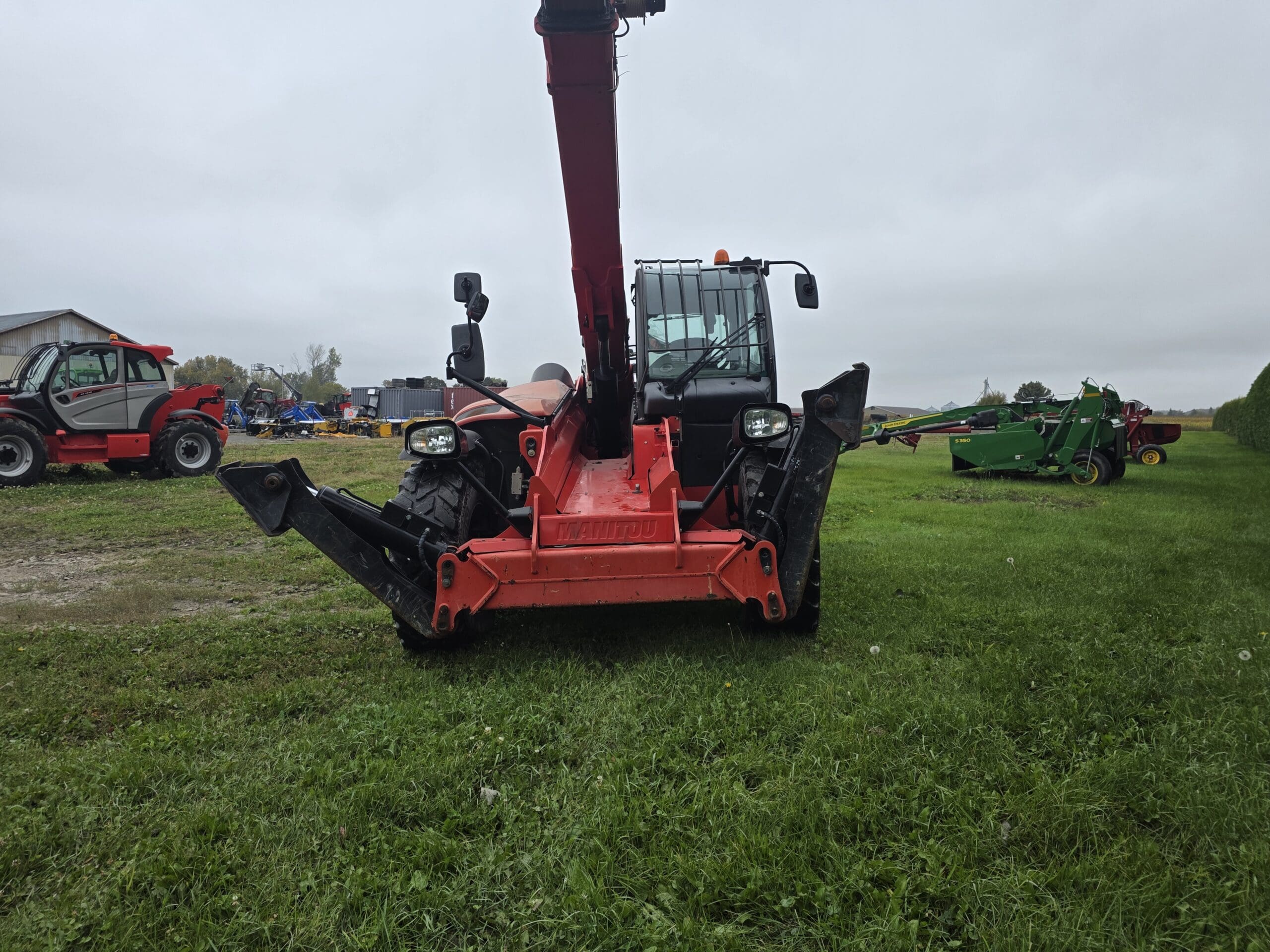 Un chariot télescopique rouge, dont la flèche est déployée, est garé sur de l'herbe verte, face à la caméra. D'autres machines agricoles et un bâtiment sont visibles à l'arrière-plan, par une journée nuageuse.