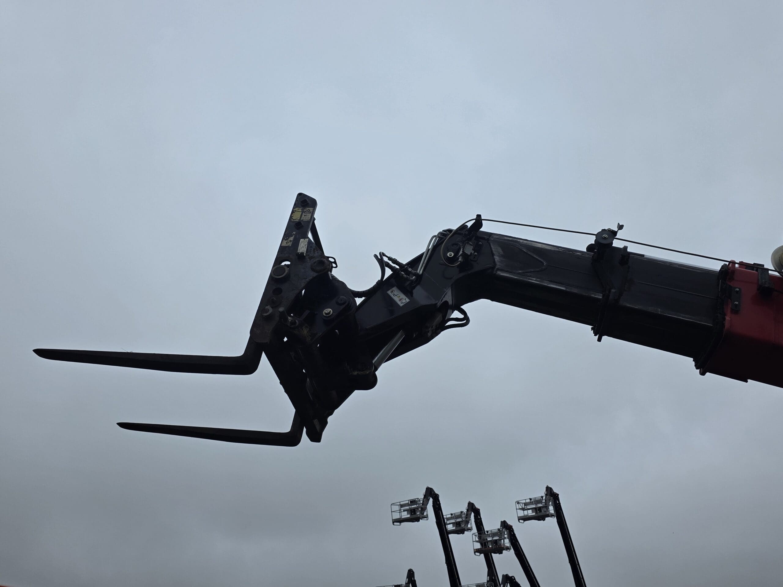 Gros plan sur un accessoire de chariot élévateur fixé au bras d'une grosse machine, se découpant sur un ciel nuageux. Plusieurs machines similaires sont visibles à l'arrière-plan.