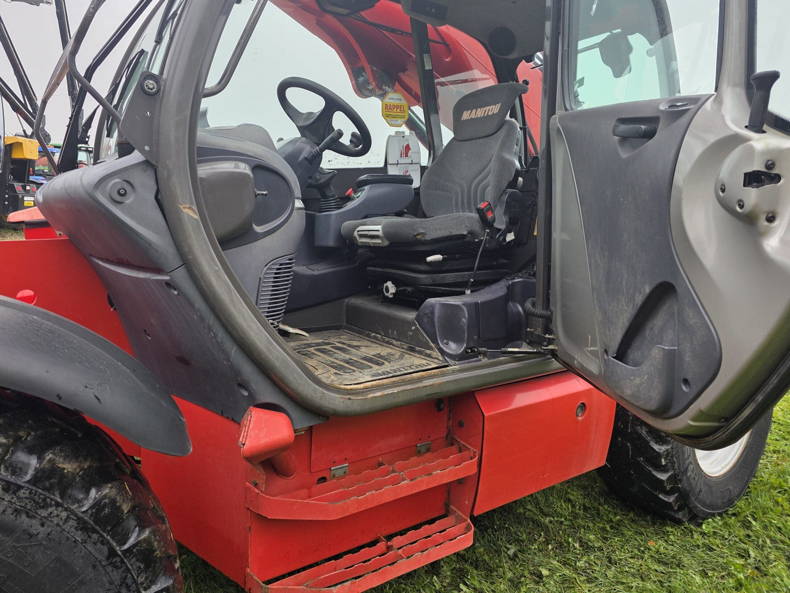 La photo montre la porte ouverte et l'intérieur de la cabine d'un véhicule utilitaire ou d'un tracteur MANITOU rouge, laissant apparaître le siège du conducteur, le volant et le marchepied recouverts de saleté. À l'extérieur, on aperçoit de l'herbe et un morceau de pneu.