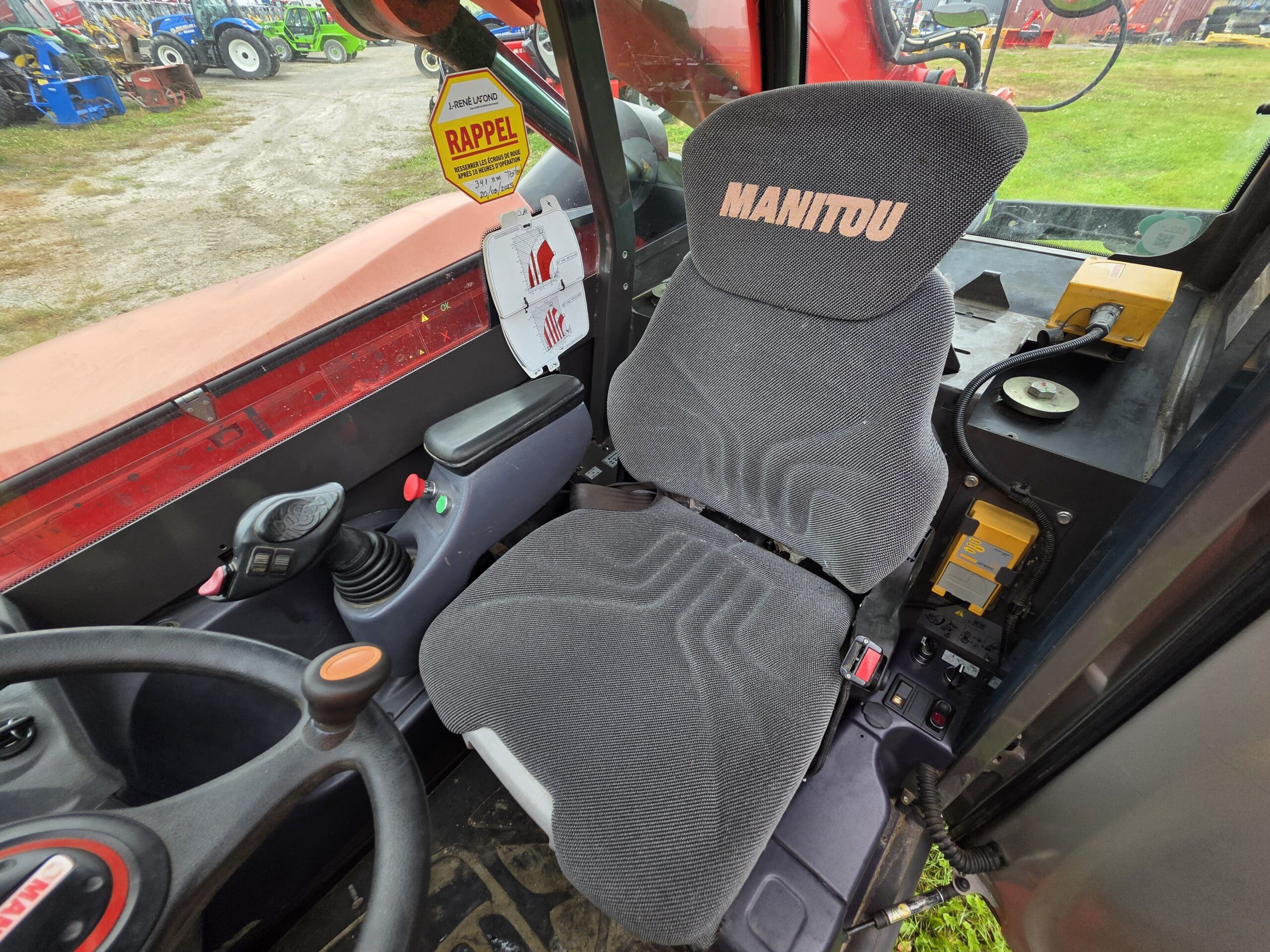 La photo montre le siège conducteur et les commandes à l'intérieur de la cabine d'un tracteur MANITOU, avec un siège rembourré gris, un volant, des leviers, des autocollants d'avertissement et divers tableaux de commande, tandis que des engins sont visibles à travers la vitre.