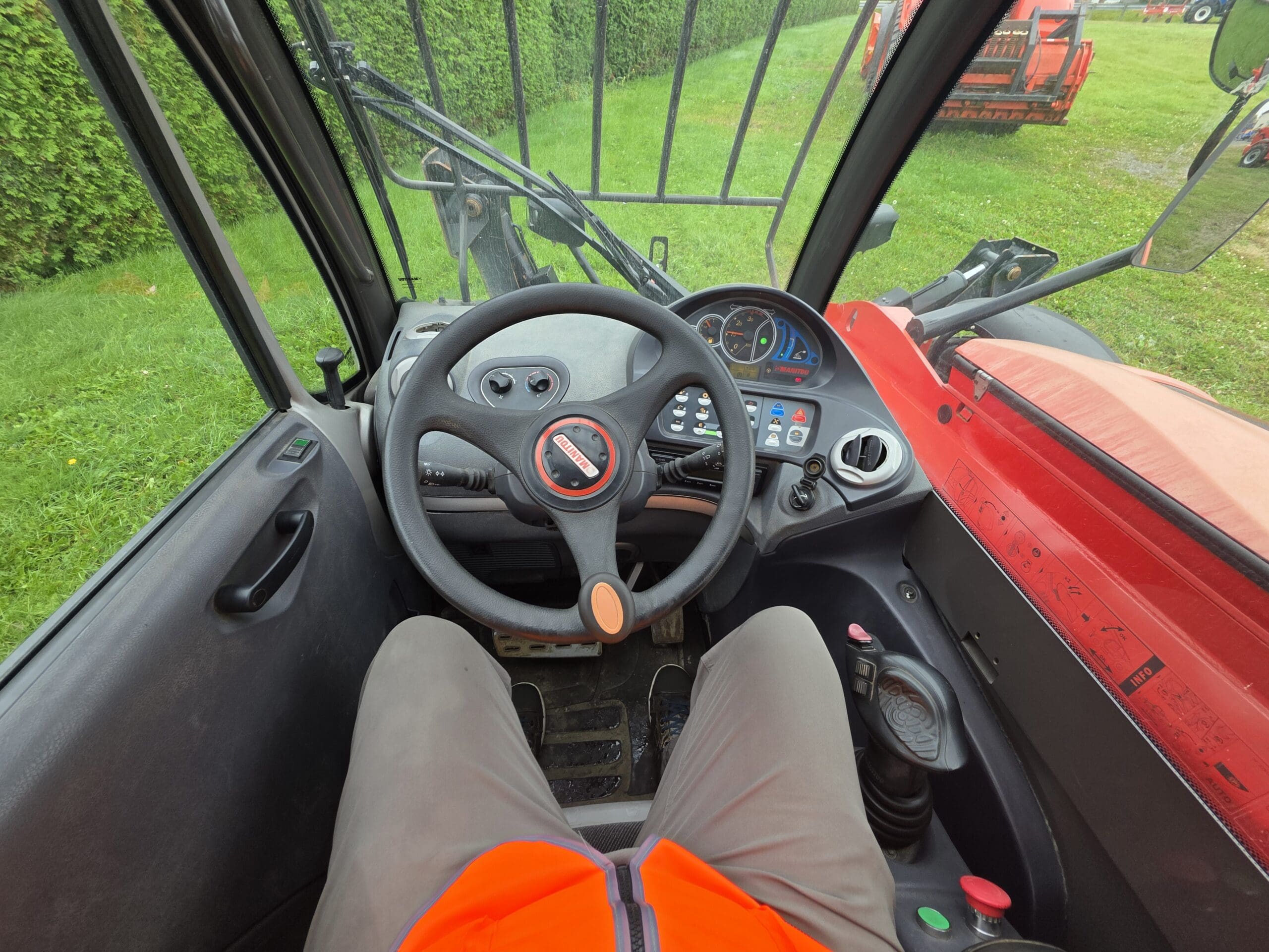 Une personne vêtue d'orange est assise au volant d'un tracteur, les mains loin du volant ; on aperçoit les tableaux de bord, les leviers et les pédales à l'intérieur de la cabine. À travers le pare-brise, on distingue de l'herbe verte et une haie.