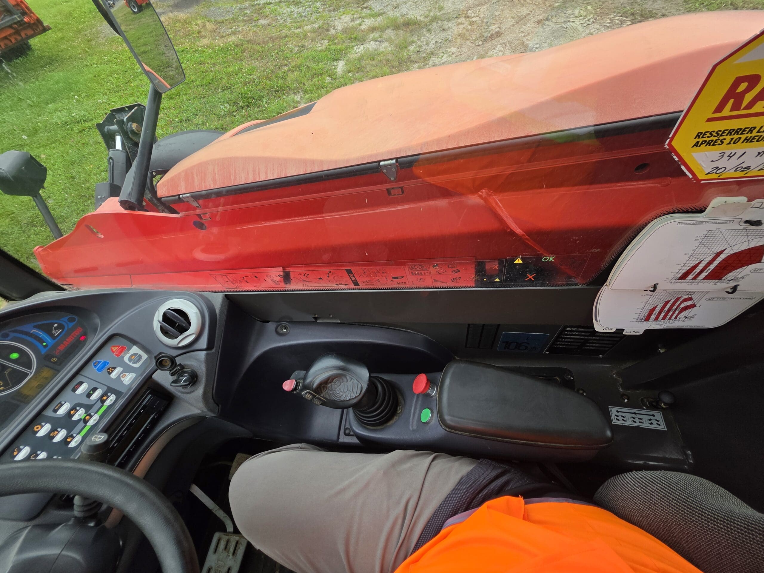 Vue depuis l'intérieur de la cabine d'un véhicule, montrant les tableaux de bord, les leviers et une personne vêtue d'un pantalon gris et d'une chemise orange, assise au volant. L'extérieur est partiellement visible à travers la vitre.