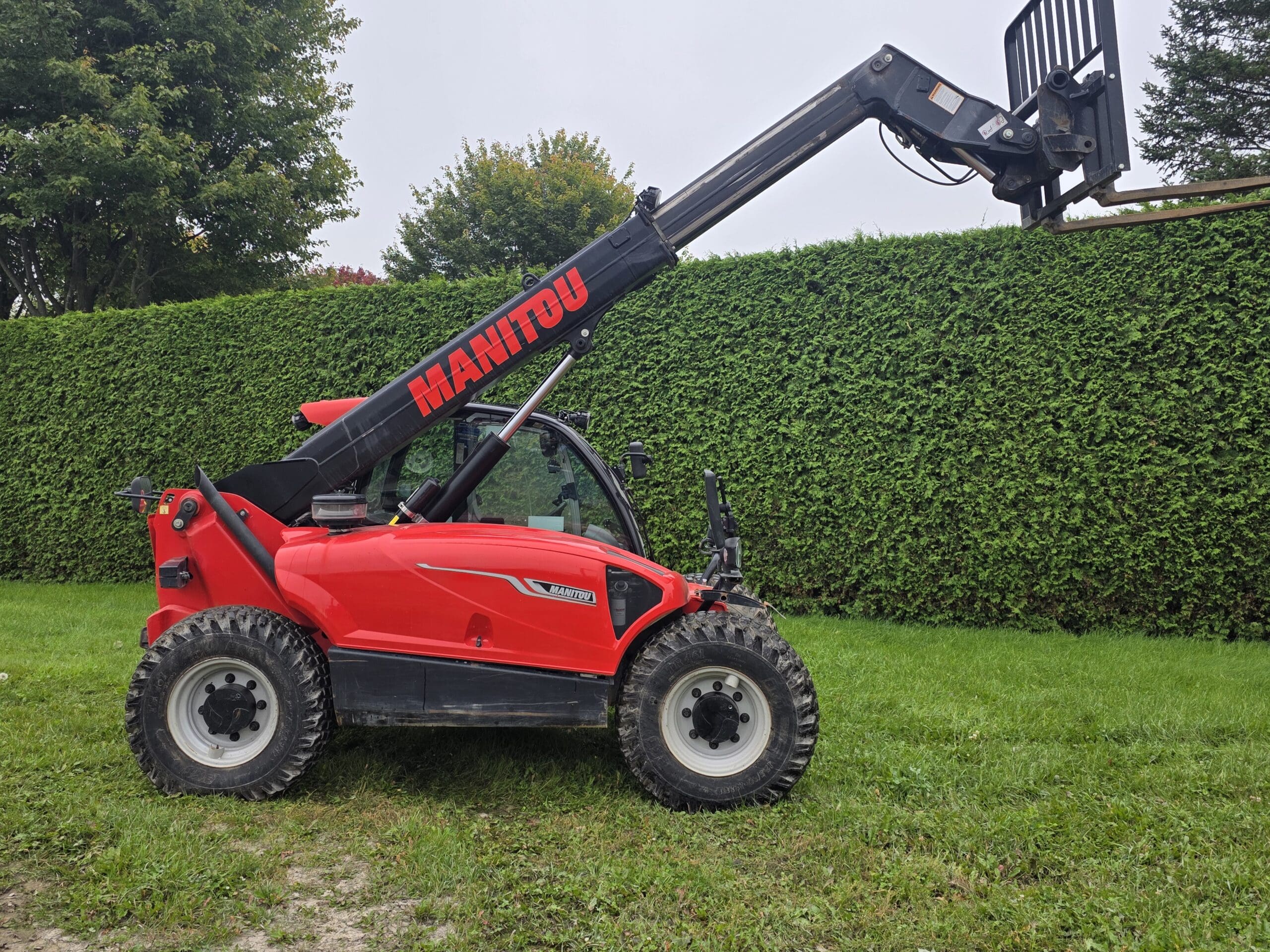 Un chariot télescopique Manitou rouge, équipé d'une flèche allongée et de fourches, est garé sur l'herbe près d'une haute haie verte et d'arbres, sous un ciel couvert.