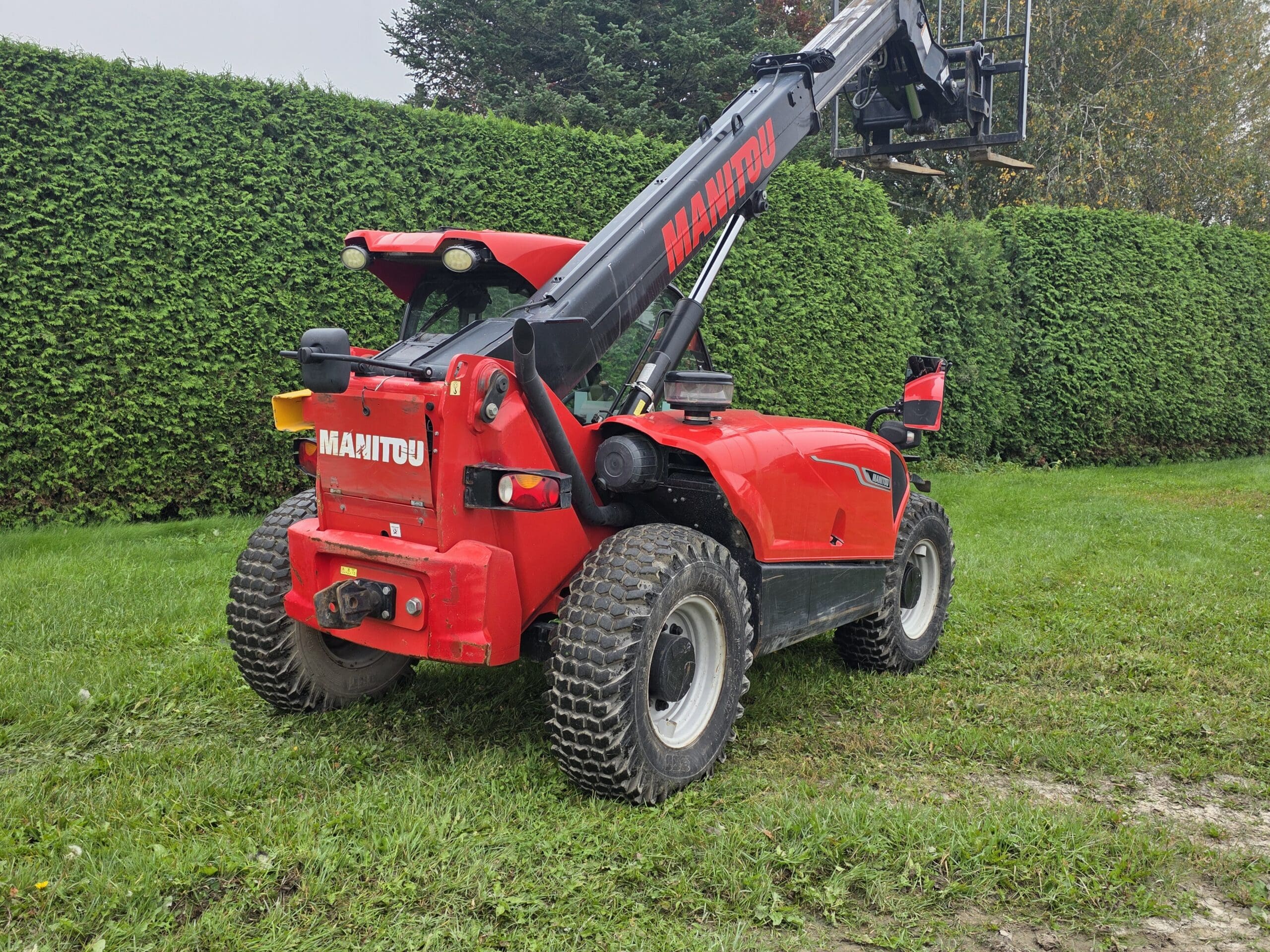 Un chariot télescopique Manitou rouge est garé sur l'herbe, près de hautes haies vertes, sa flèche étant partiellement relevée. La scène se déroule en extérieur, par une journée nuageuse.