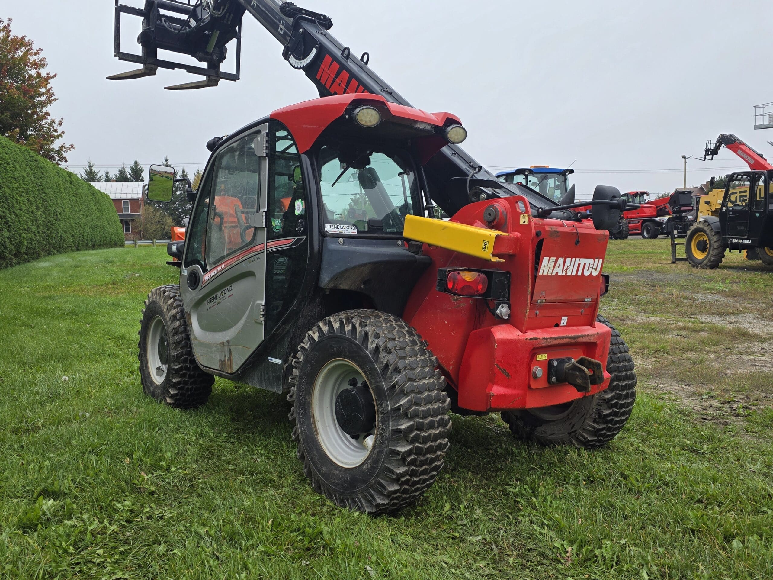 Un chariot élévateur télescopique Manitou rouge est garé sur l'herbe, le bras levé. Plusieurs autres engins de chantier sont visibles à l'arrière-plan, sous un ciel couvert.