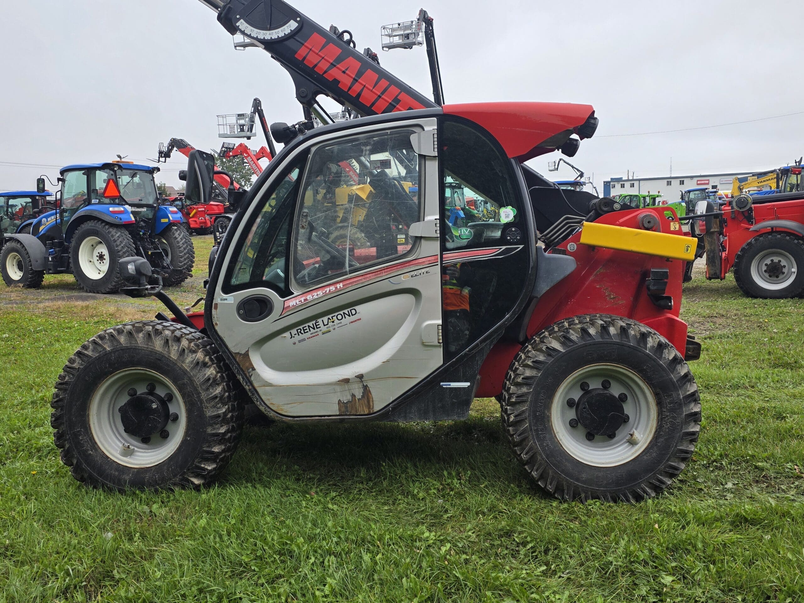 Un chariot télescopique rouge et blanc équipé de gros pneus est garé sur l'herbe, au milieu d'autres machines agricoles, notamment des tracteurs bleus, par une journée nuageuse.