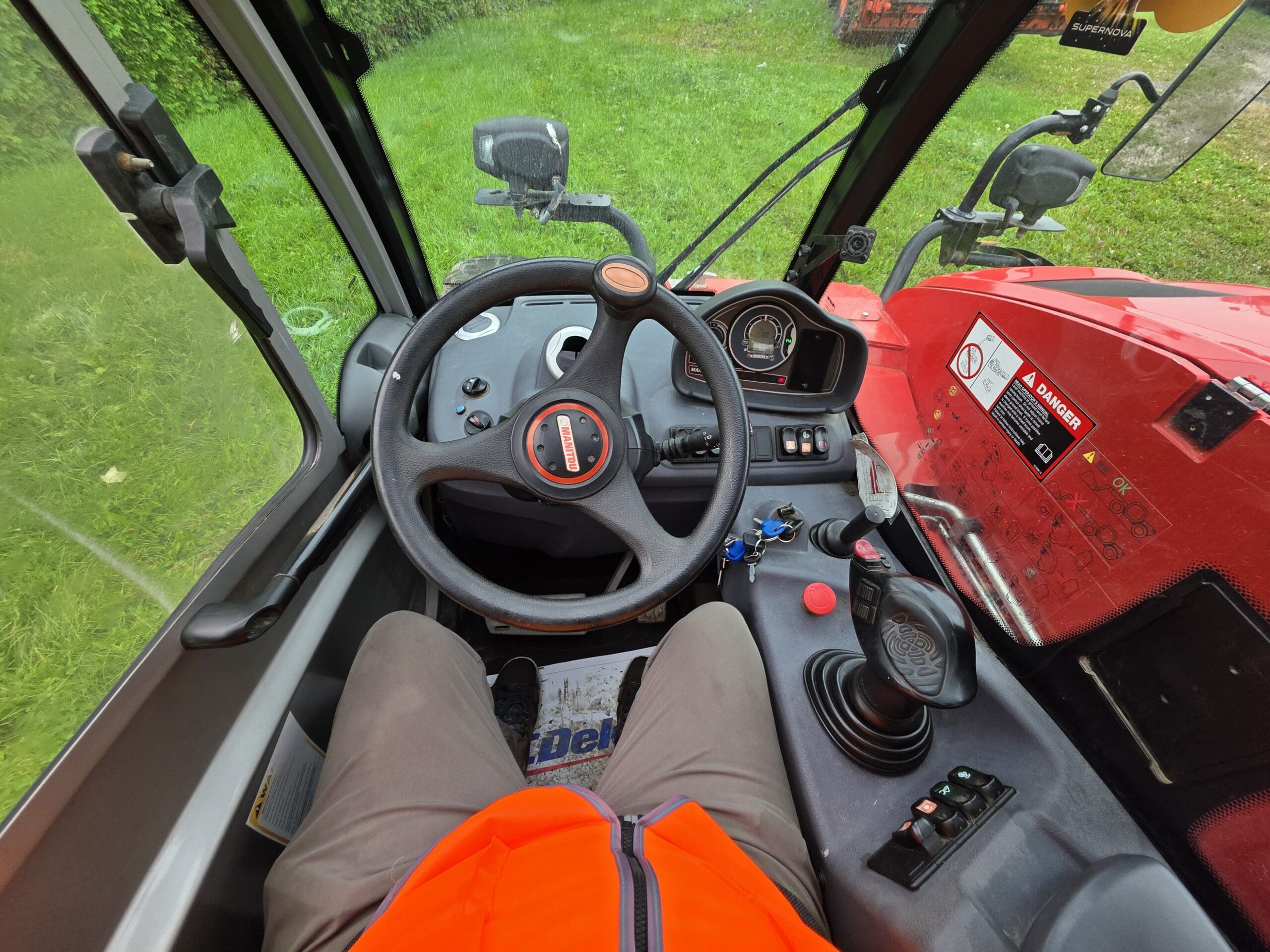 Vue depuis l'intérieur de la cabine d'un tracteur rouge, montrant le volant, le tableau de bord, le levier de vitesses, les commutateurs, ainsi qu'une personne vêtue d'un pantalon gris et d'un gilet orange, assise au poste de conduite. On aperçoit de l'herbe à l'extérieur.
