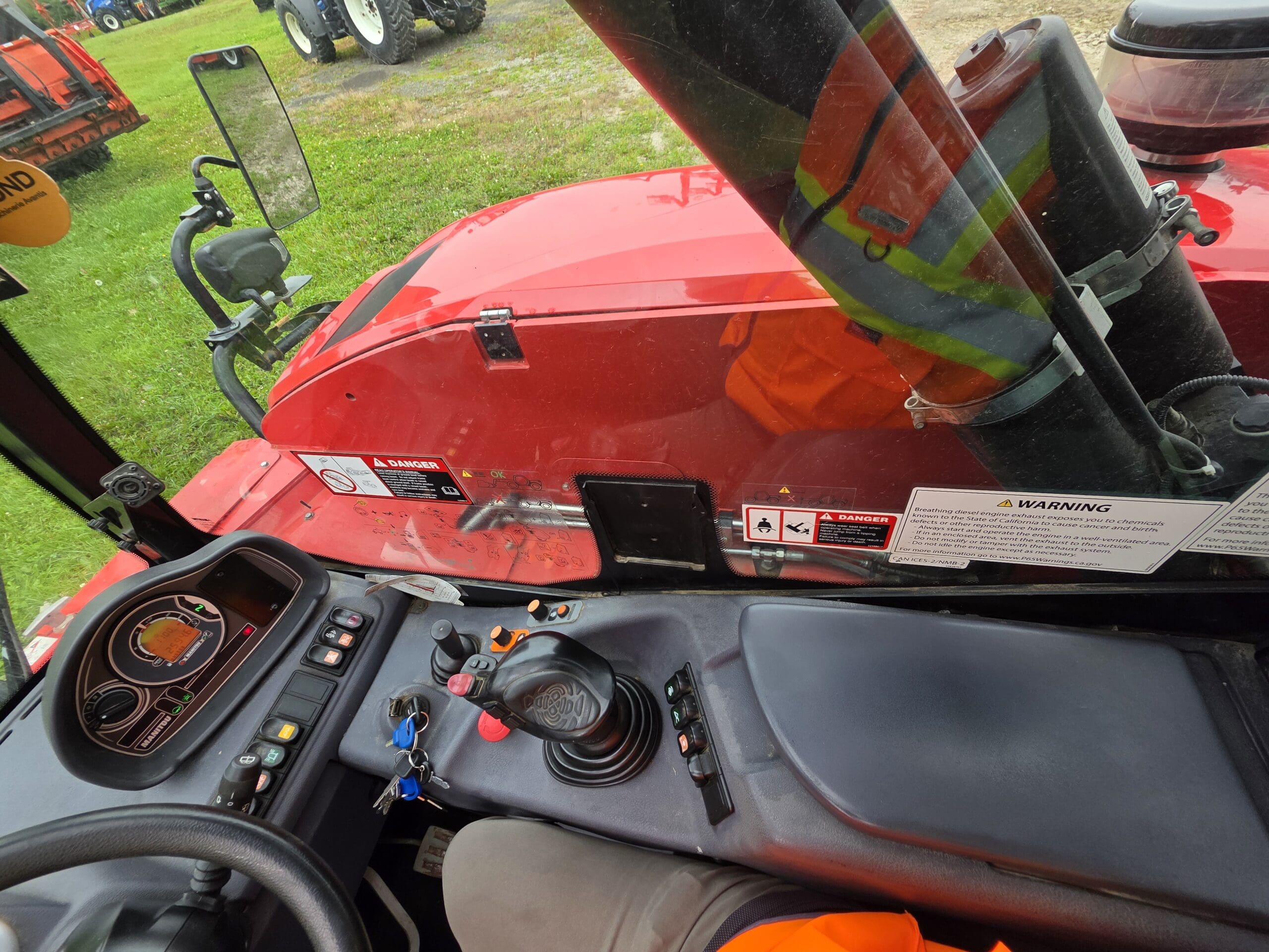 Vue depuis l'intérieur de la cabine d'un tracteur rouge, montrant le tableau de bord, le joystick, les étiquettes d'avertissement et une personne portant un gilet de sécurité orange, avec à l'extérieur, à travers les vitres, un sol recouvert d'herbe.