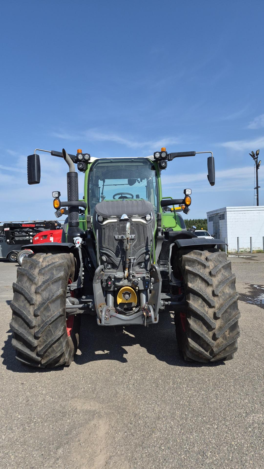 Un tracteur vert équipé de gros pneus robustes est garé sur la chaussée sous un ciel bleu limpide ; on le voit de face. D'autres véhicules et un bâtiment blanc apparaissent à l'arrière-plan.