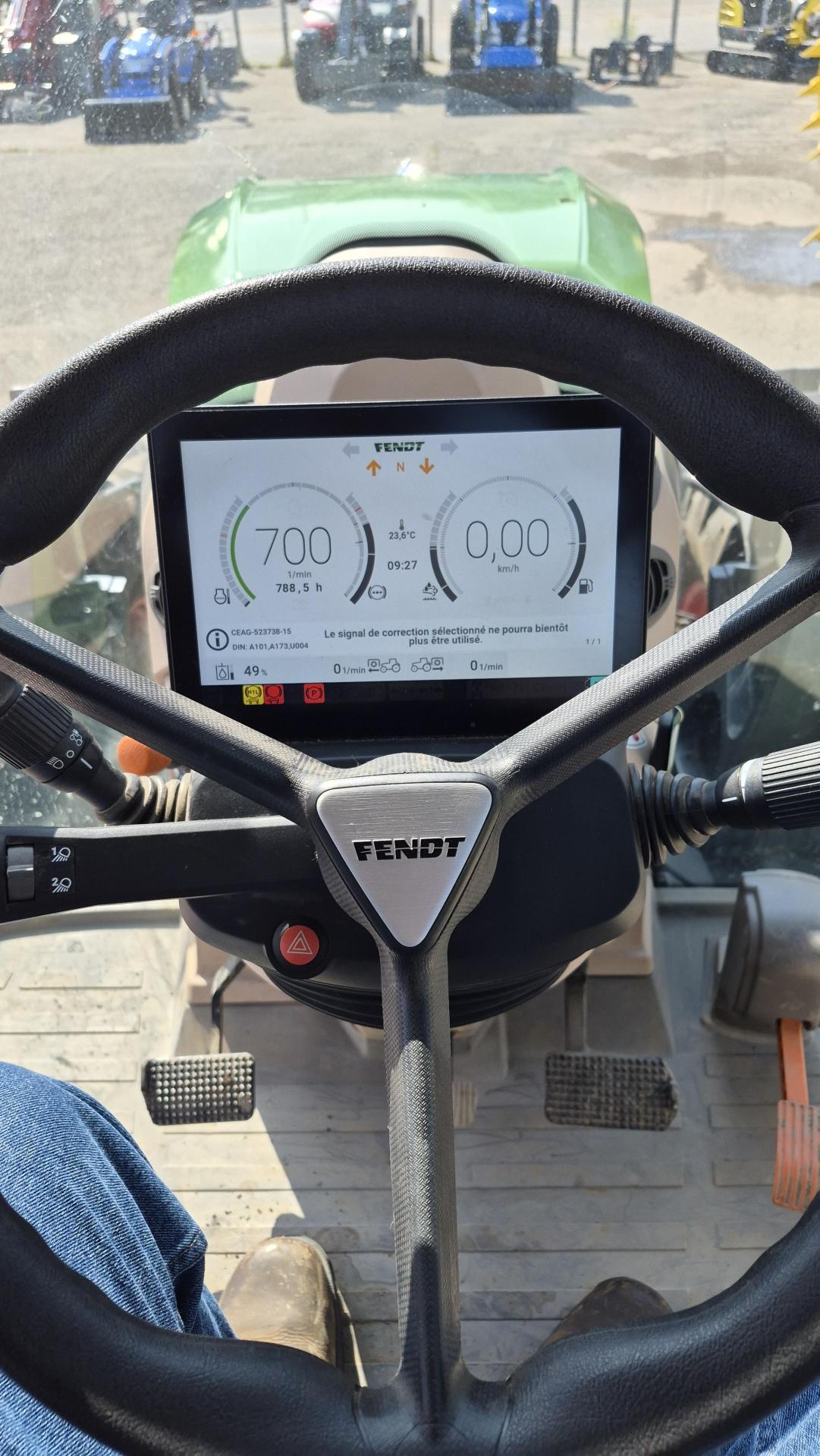 Vue depuis le siège conducteur d'un tracteur Fendt, montrant le volant et l'écran du tableau de bord affichant la vitesse et les données du moteur. Des boutons et des commandes sont visibles autour du siège.