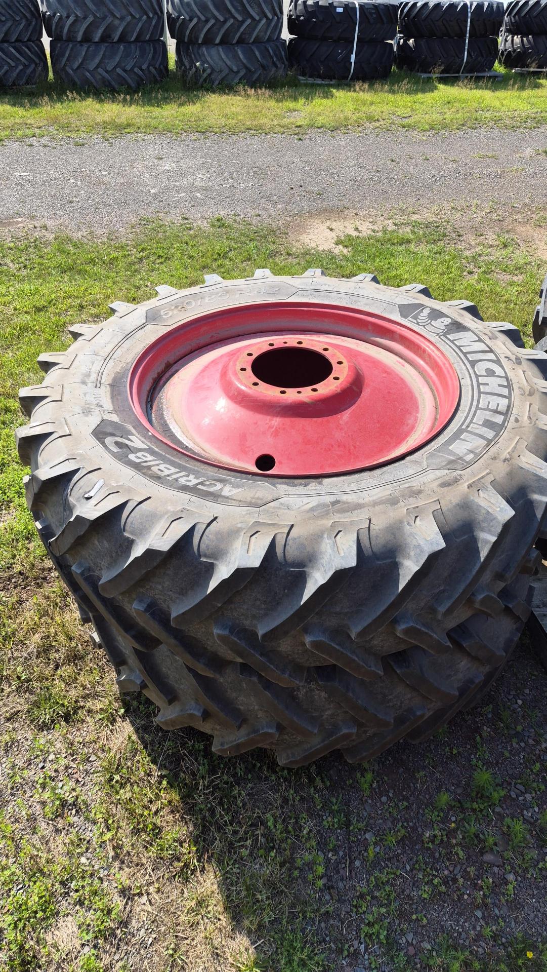 Un gros pneu de tracteur, dont la jante est rouge, repose sur de l'herbe et du gravier. D'autres pneus empilés sont visibles à l'arrière-plan. Le pneu présente des rainures profondes et la marque « Michelin » est visible sur le flanc.