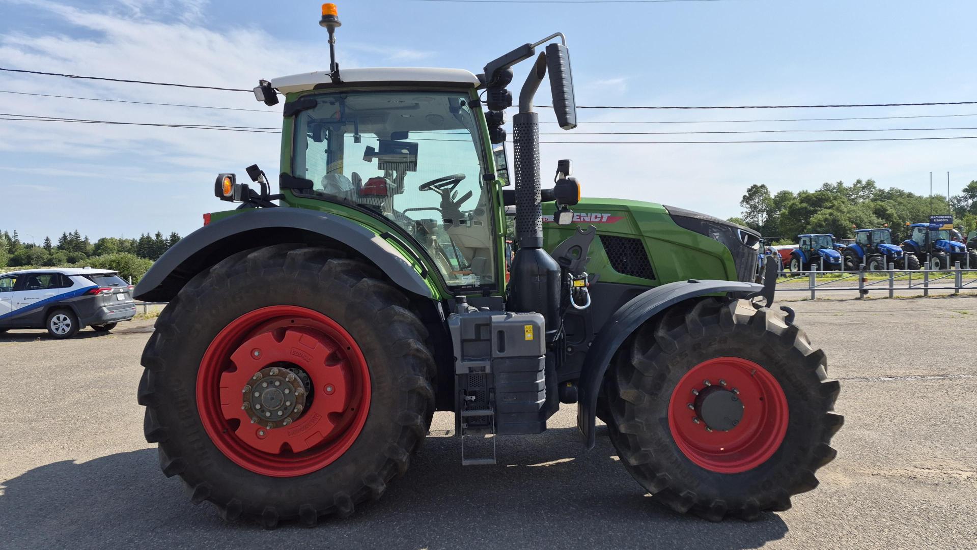 Un tracteur vert équipé de grandes roues rouges est garé sur une aire pavée par une journée ensoleillée. D'autres véhicules et tracteurs sont visibles à l'arrière-plan.