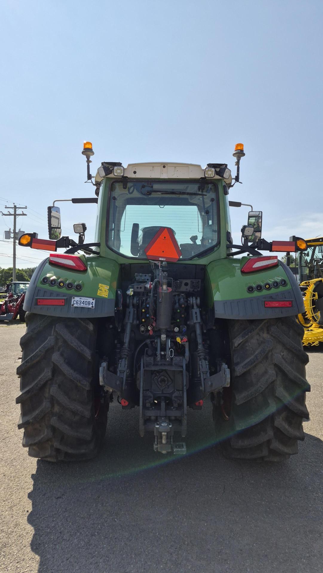 Un tracteur vert vu de dos, doté de gros pneus noirs, de feux de signalisation, d'un panneau triangulaire rouge signalant un véhicule lent, ainsi que de divers composants mécaniques visibles sous un ciel dégagé.