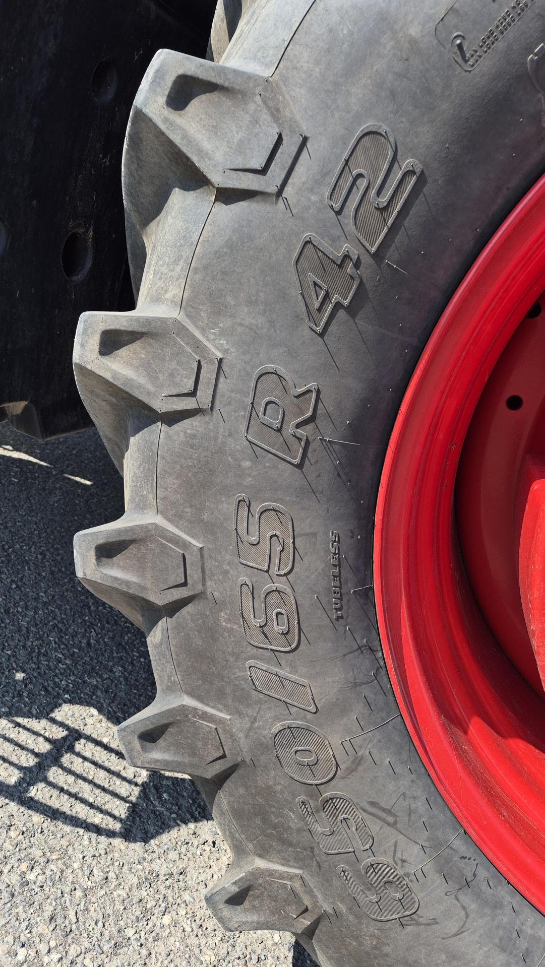 Gros plan sur un grand pneu noir de tracteur, doté de rainures profondes et portant l'inscription « 650/65 R 42 ». La jante est peinte en rouge vif. La lumière du soleil projette des ombres sur la chaussée à côté du pneu.