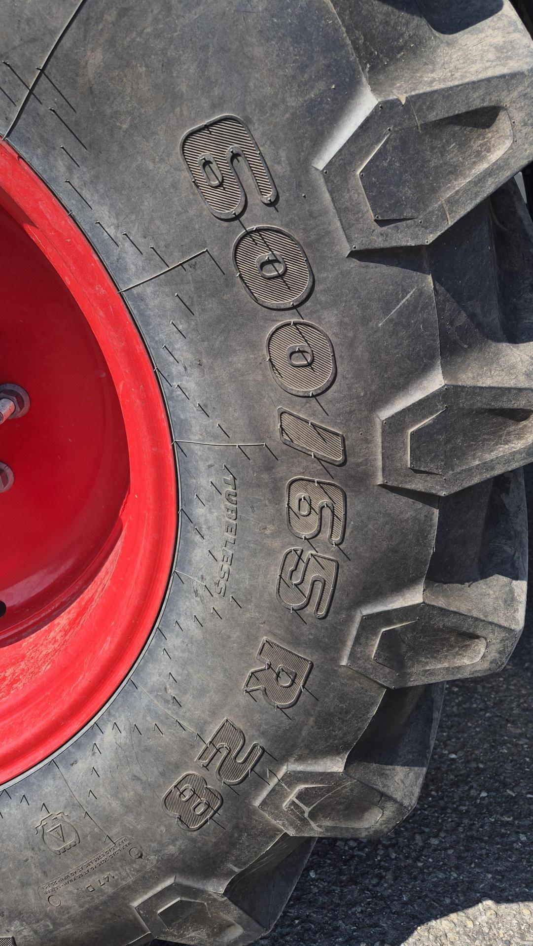 Gros plan sur un grand pneu noir de tracteur, doté de rainures profondes et d’une jante rouge vif. Les indications de dimension du pneu « 600/65 R 38 » sont visibles sur le caoutchouc. Le pneu se trouve sur une surface extérieure revêtue.