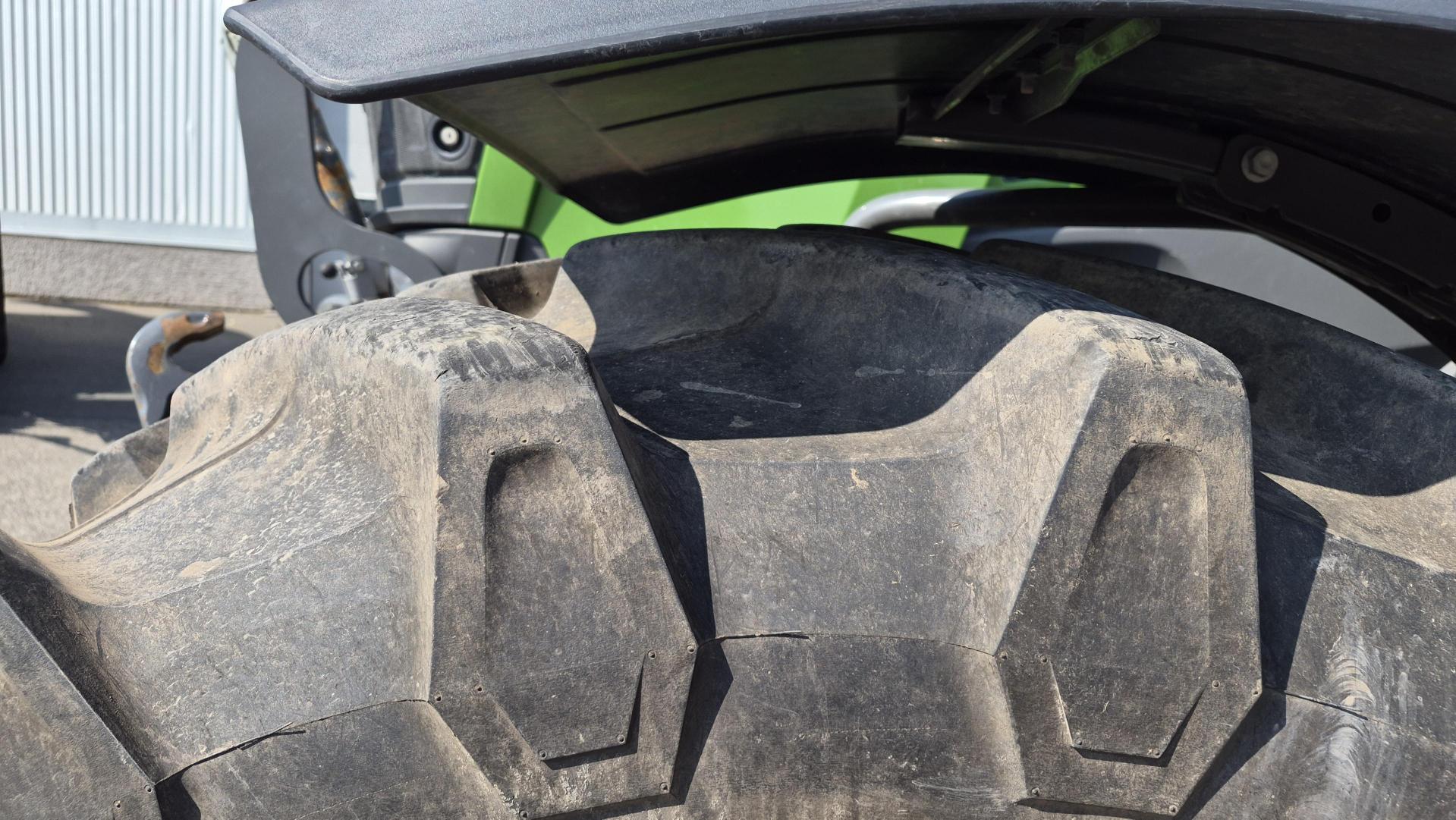 Gros plan sur un gros pneu de tracteur poussiéreux, aux rainures profondes, partiellement recouvert par l'aile du véhicule. Une partie de la carrosserie verte du tracteur et un bout de chaussée sont visibles à l'arrière-plan.