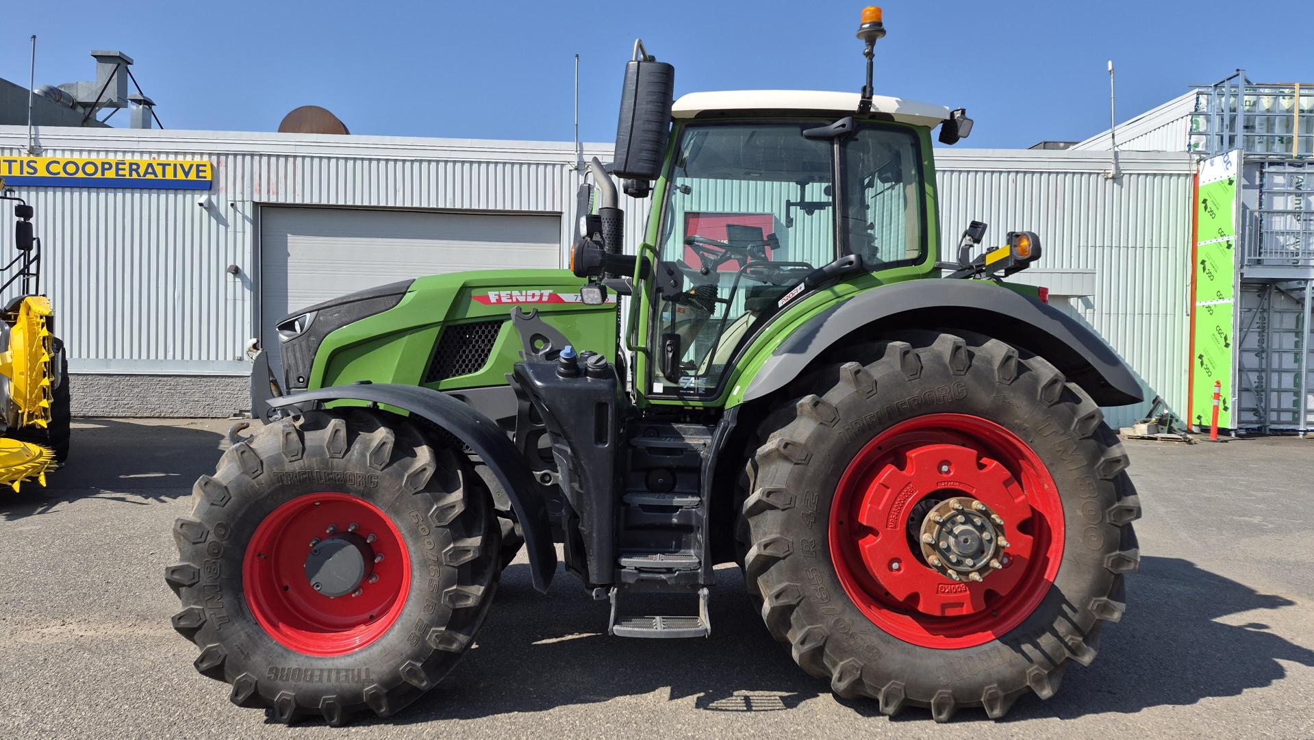 Un tracteur Fendt vert, équipé de grandes roues rouges, est garé sur le trottoir devant un bâtiment blanc portant l'inscription « COOPERATIVE », sous un ciel d'un bleu limpide.