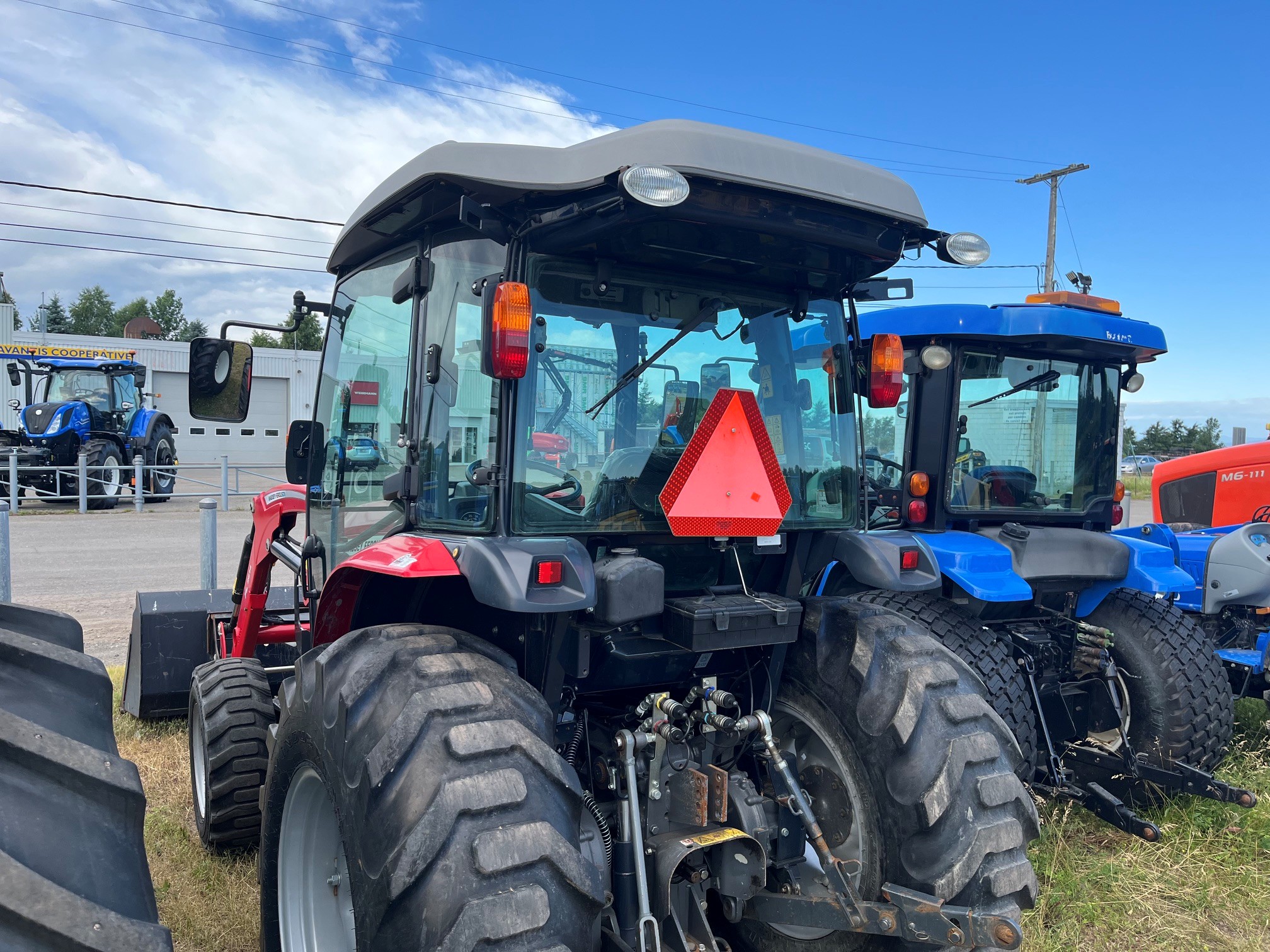Un tracteur rouge est garé sur l'herbe à côté d'un tracteur bleu ; on les voit tous les deux de dos. Un triangle réfléchissant orange vif est fixé sur le tracteur rouge. D'autres tracteurs et un ciel nuageux sont visibles à l'arrière-plan.