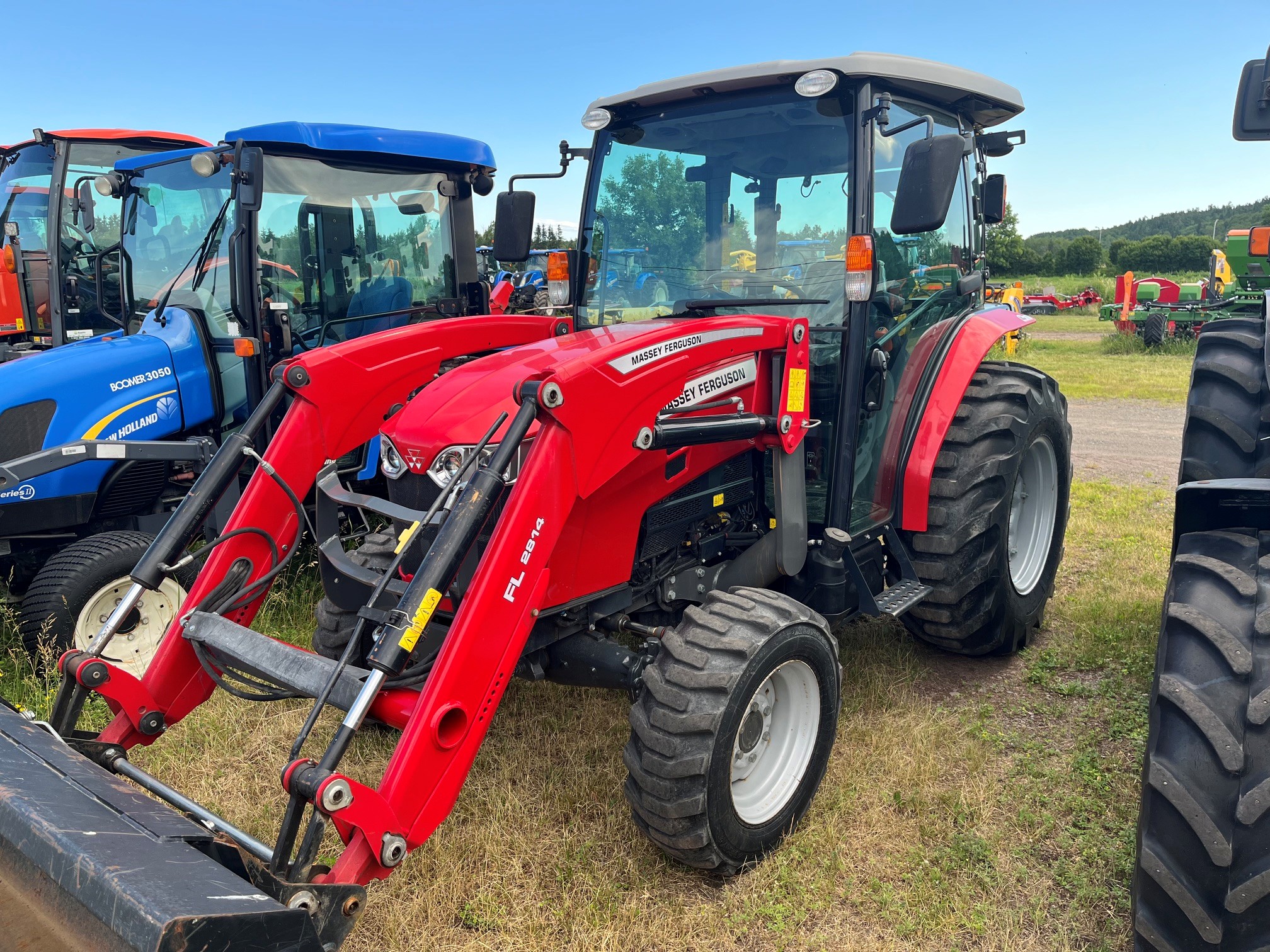 Un tracteur Massey Ferguson rouge équipé d'un chargeur frontal est garé sur l'herbe, à côté d'autres tracteurs, sous un ciel bleu. On aperçoit des arbres et d'autres machines agricoles à l'arrière-plan.