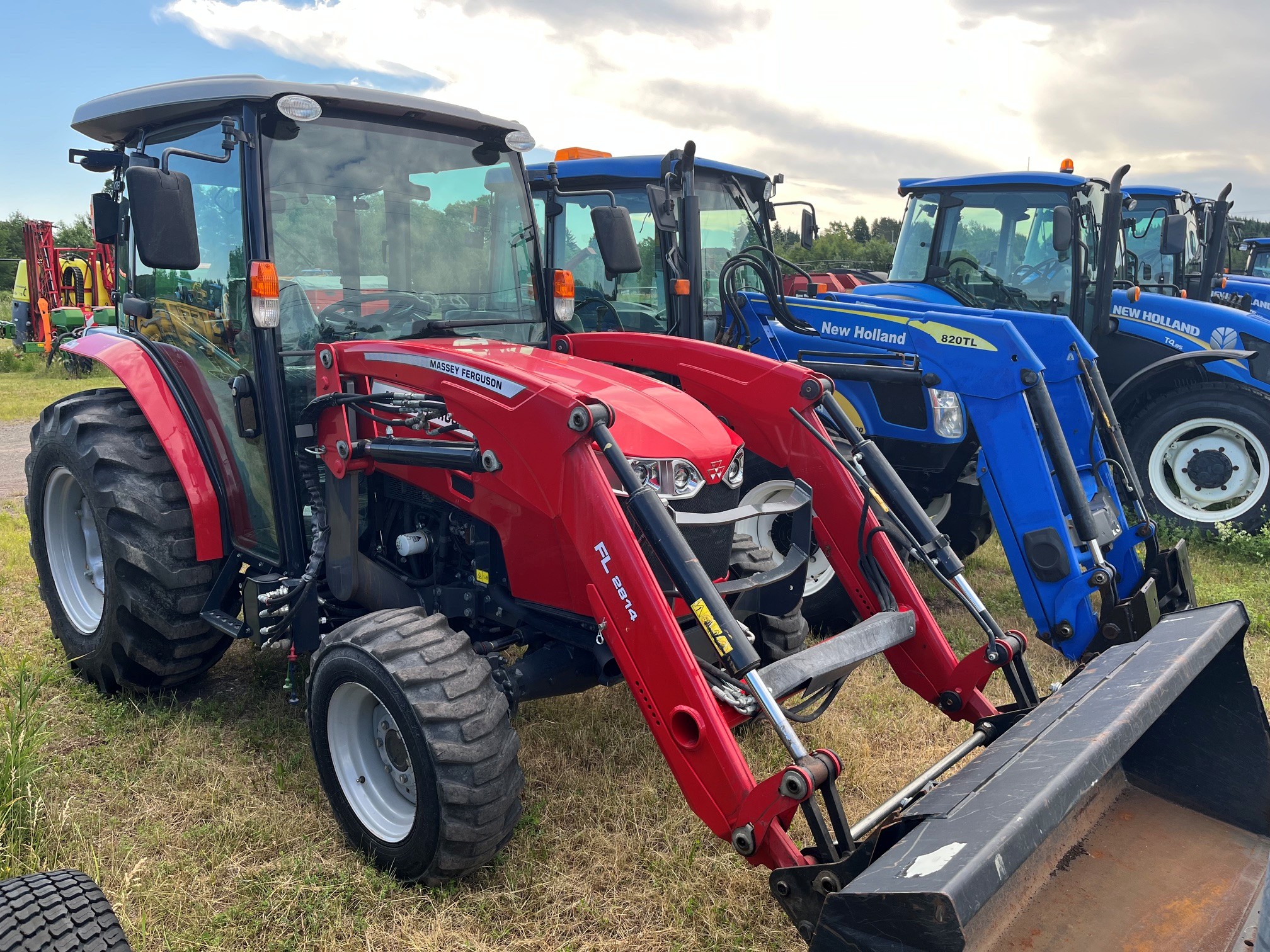 Un tracteur Massey Ferguson rouge équipé d'un chargeur frontal est garé à côté d'un tracteur New Holland bleu sur un terrain herbeux ; d'autres tracteurs sont visibles à l'arrière-plan, sous un ciel nuageux.