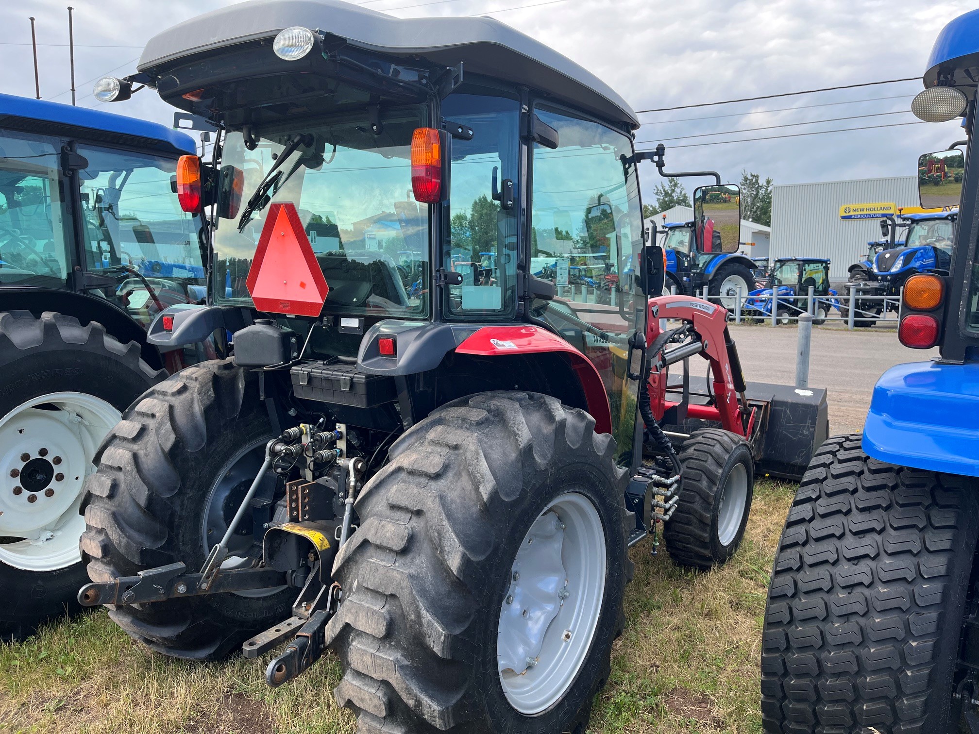 Un tracteur rouge et noir équipé d'une cabine est garé sur l'herbe, au milieu d'autres tracteurs, dans un parc extérieur ; on aperçoit en arrière-plan un ciel nuageux et les enseignes du concessionnaire.