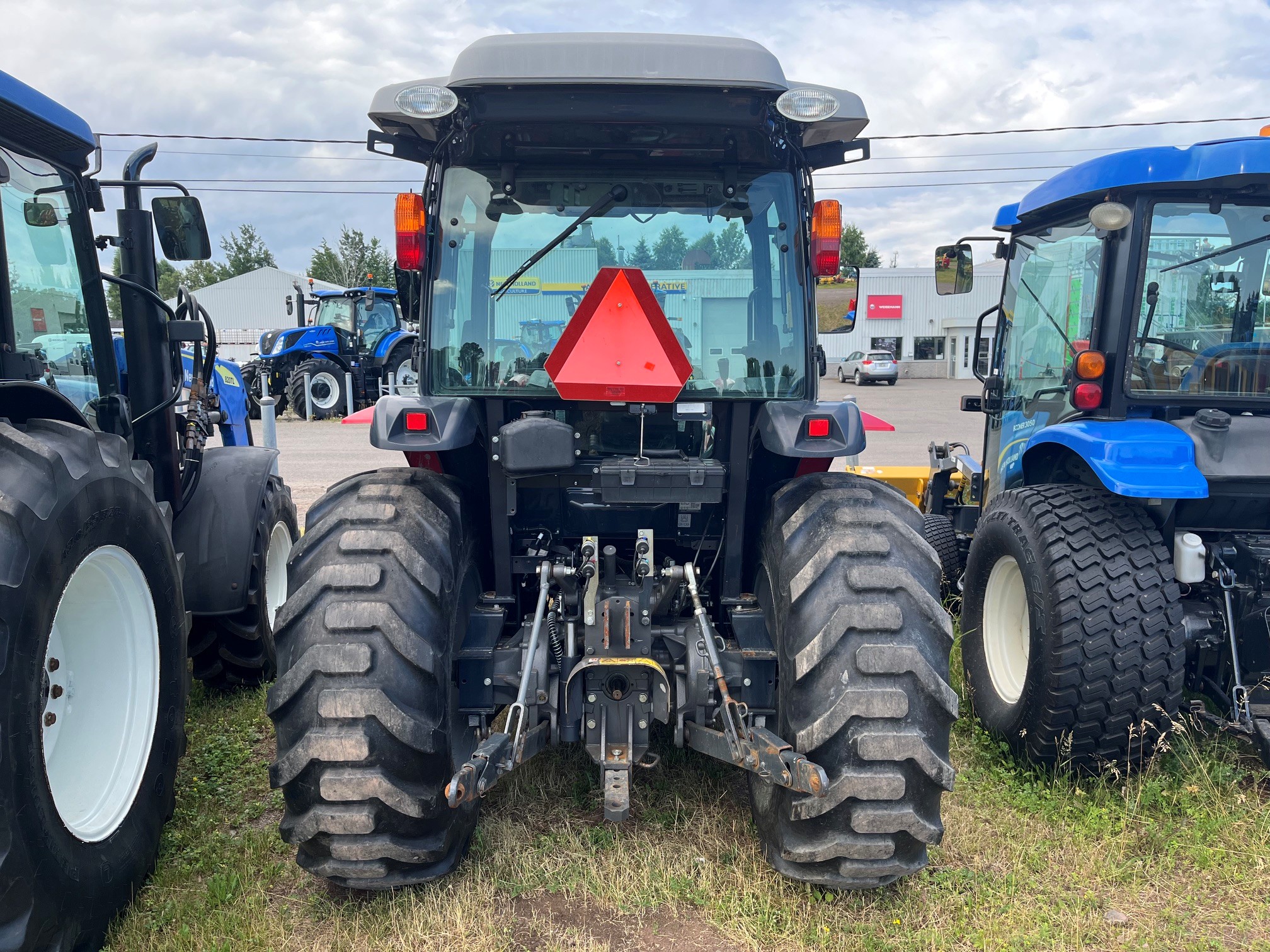 Vue arrière d'un tracteur agricole noir garé à l'extérieur entre deux tracteurs bleus. On remarque ses gros pneus à crampons profonds, un panneau triangulaire rouge central signalant un véhicule lent, ainsi que des articulations mécaniques visibles dans la partie inférieure.